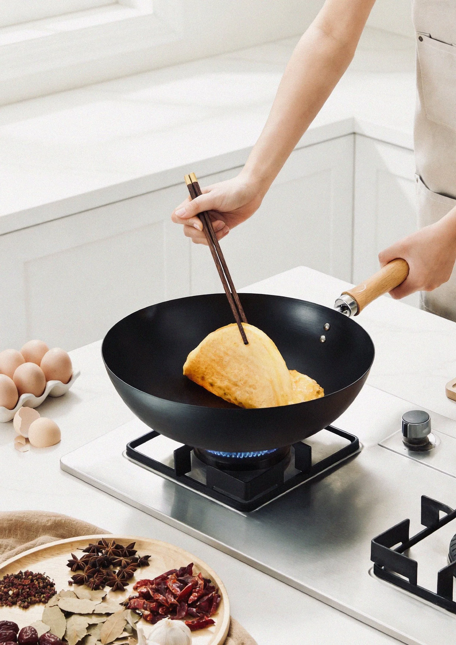 A person cooking an omelette in a black non-stick skillet on a stovetop. The person is holding chopsticks with one hand and the handle of the skillet with the other. There are eggs and a bowl nearby, along with various spices on a wooden tray.