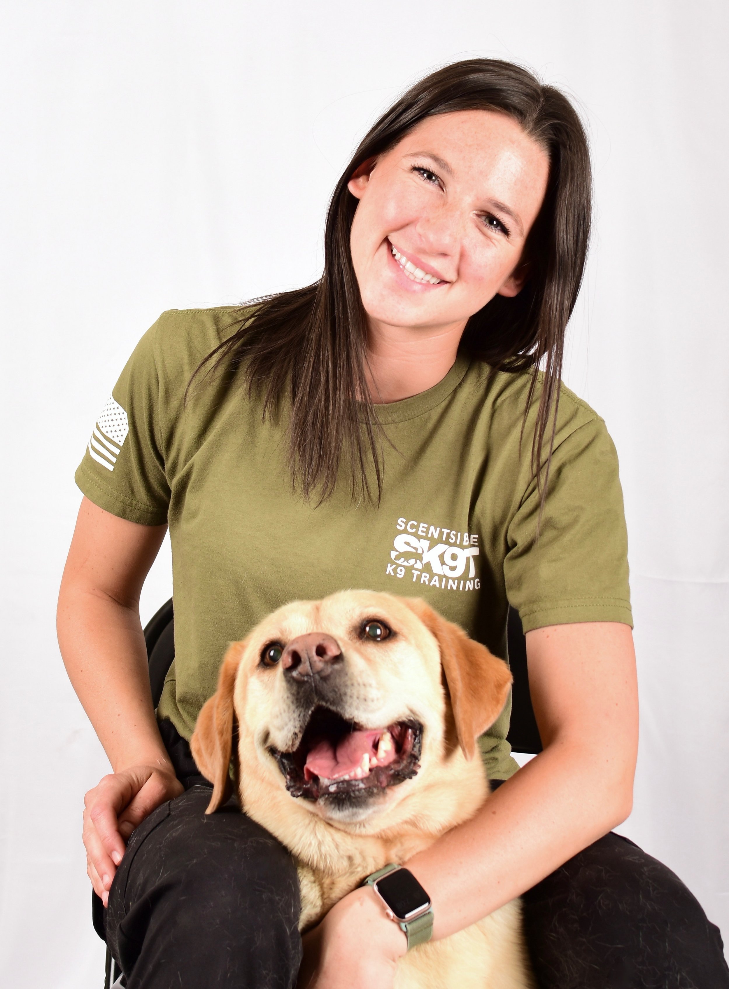 A woman with long brown hair smiling and sitting with a yellow dog on her lap.
