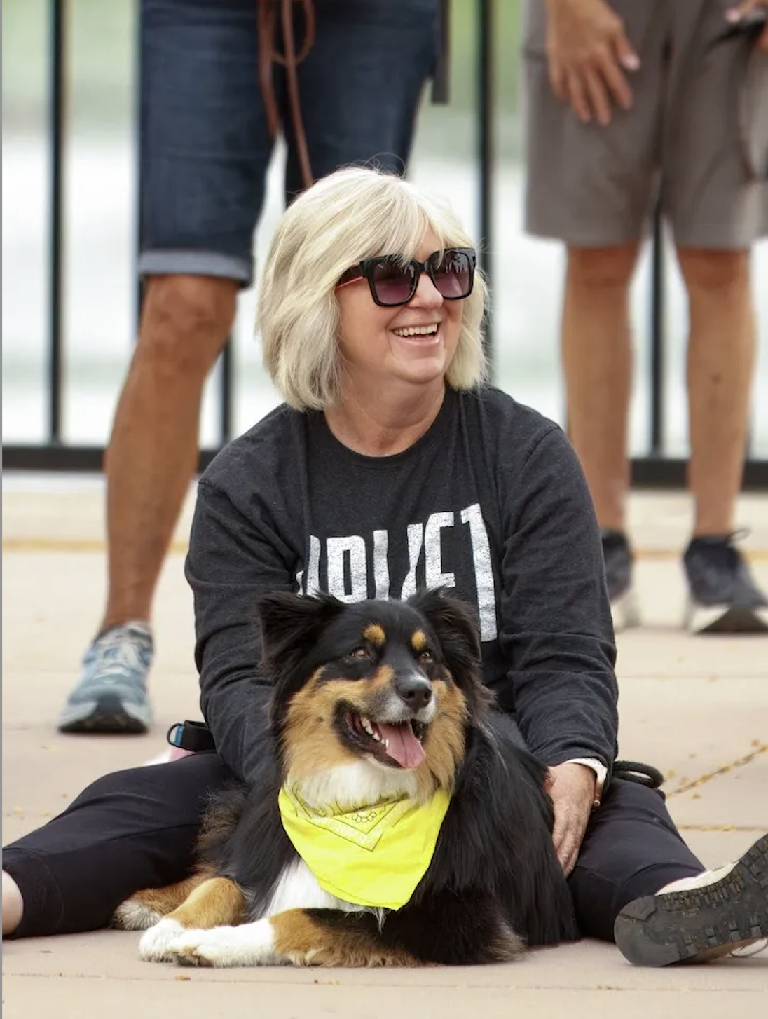 A woman with blonde hair wearing sunglasses and a black shirt with the word 'POLICE' written on it, sitting on the ground smiling and holding a black and tan Australian Shepherd dog with a yellow bandana, in an outdoor setting with people standing in the background.