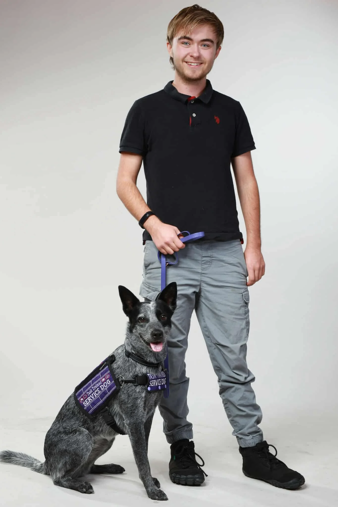 Young man in black polo shirt and gray cargo pants holding a blue leash attached to a service dog with a purple vest, sitting on a white background.