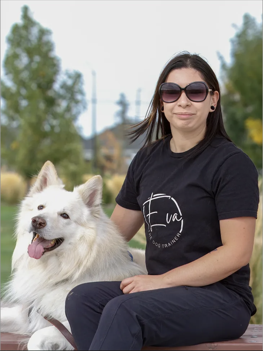 A woman with black hair and sunglasses sitting next to a white fluffy dog with a pink tongue hanging out outdoors during daytime.