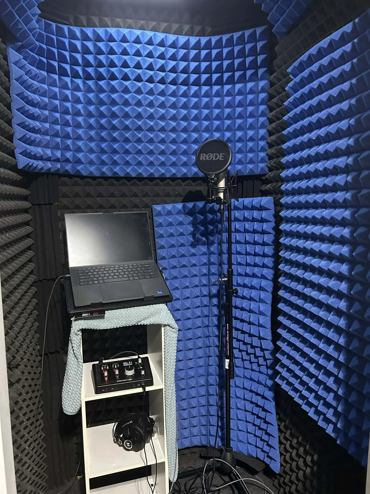 Recording studio with blue and black soundproof foam panels, a laptop on a small table, a microphone with a pop filter, a sound mixer, and headphones hanging on a shelf.
