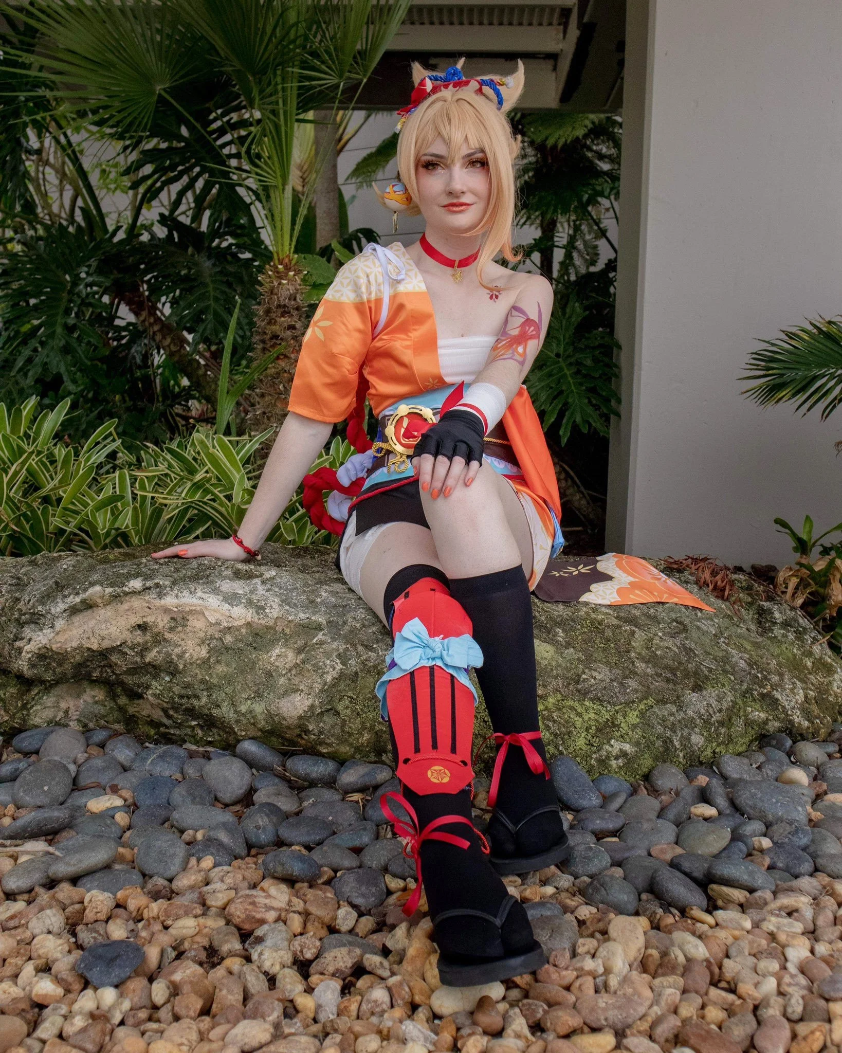 Person in a colorful anime-inspired costume sitting on a large rock outdoors surrounded by tropical plants and pebbles.