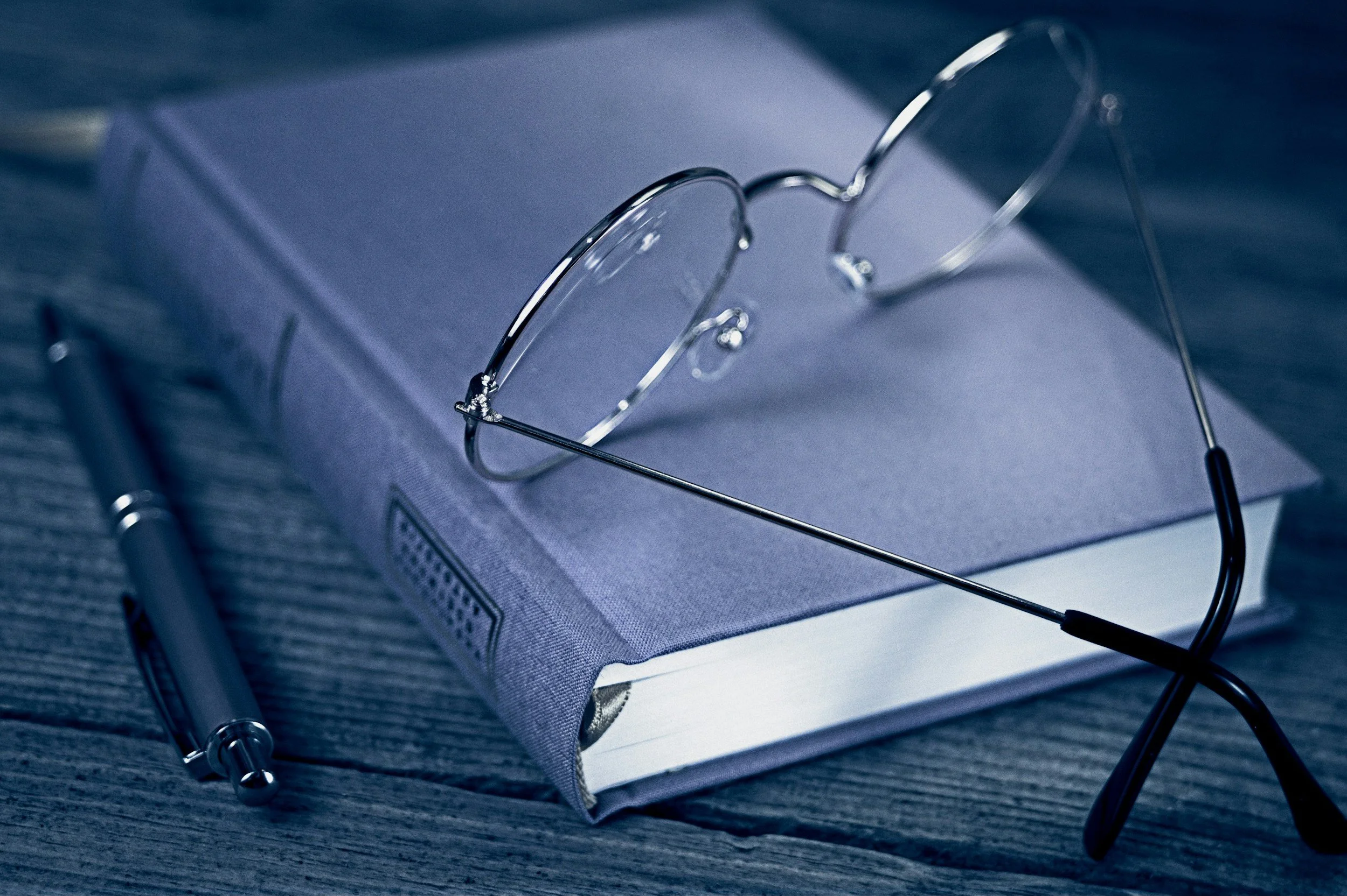 A pair of reading glasses resting on a closed, purple hardcover book on a wooden surface, accompanied by a silver pen.