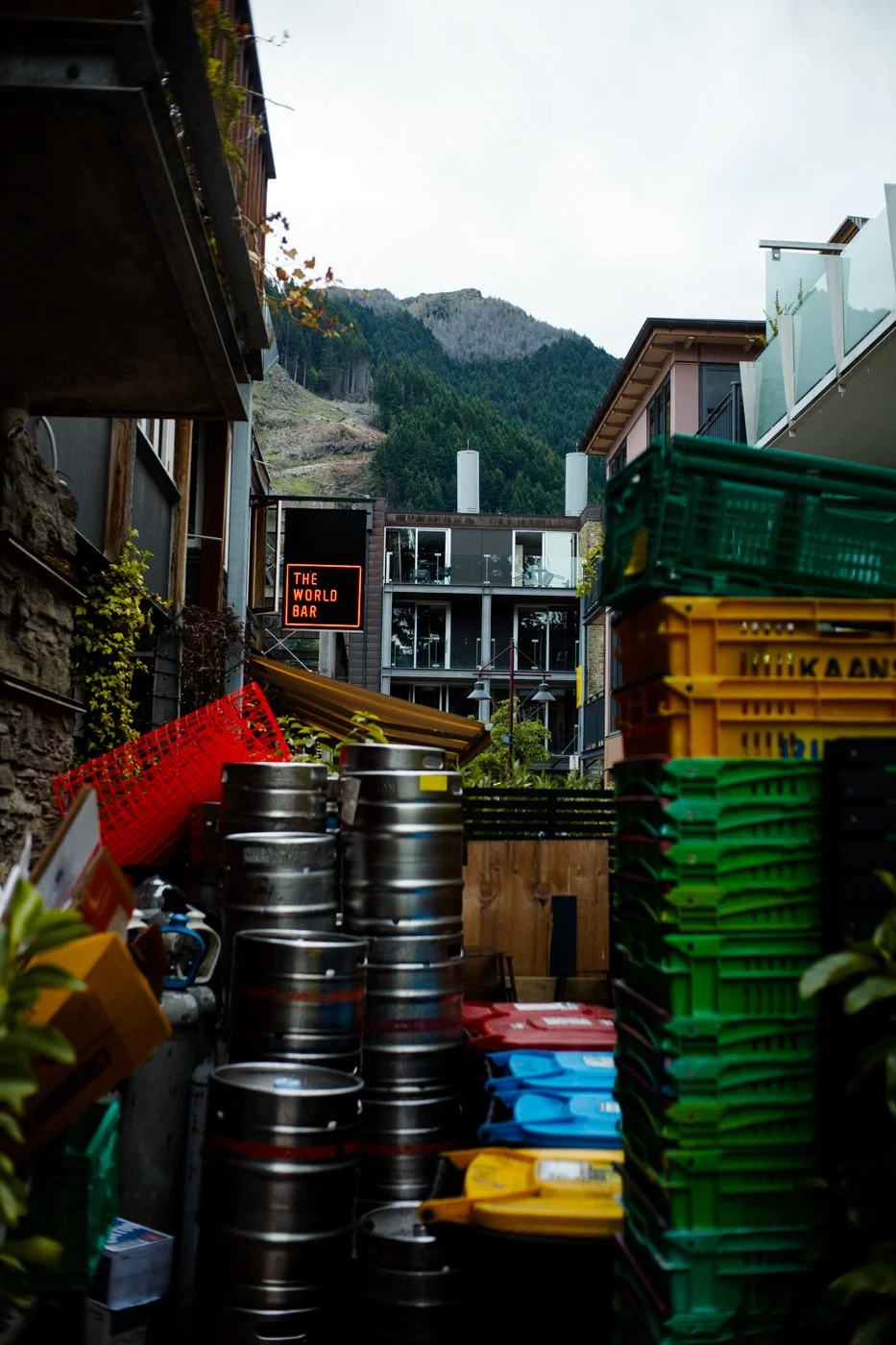 Alleyway scene with stacked colored crates and metal barrels, a sign reading 'The World Bar', in a mountain town setting with modern buildings and green mountains in the background.