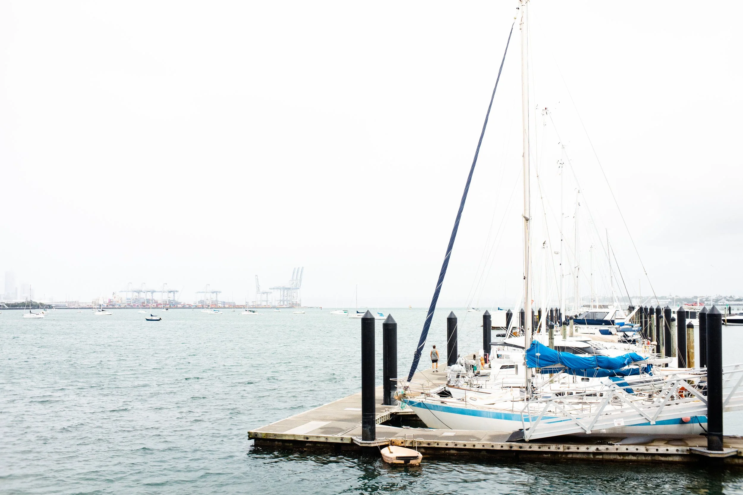 Yachts docked at a marina with a city skyline and harbor cranes in the background.