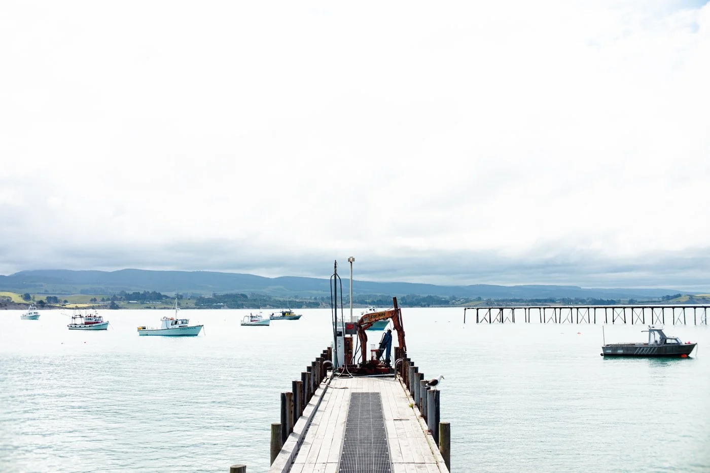 A wooden pier extends into a calm body of water with several boats anchored nearby, and rolling hills with a cloudy sky in the background.