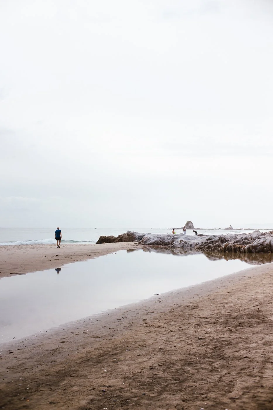 People walking on a sandy beach with rocks and a small pool of water reflecting the sky, overcast sky with the ocean in the background.