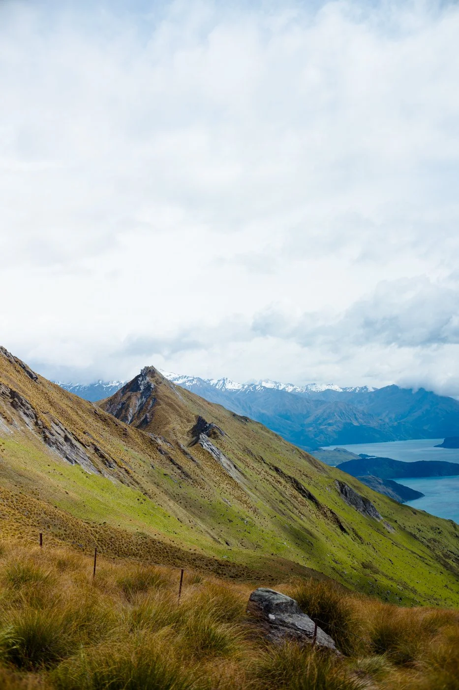 Scenic mountain landscape with grassy slopes, jagged rocks, distant snow-capped peaks, and a lake in the background under cloudy sky.