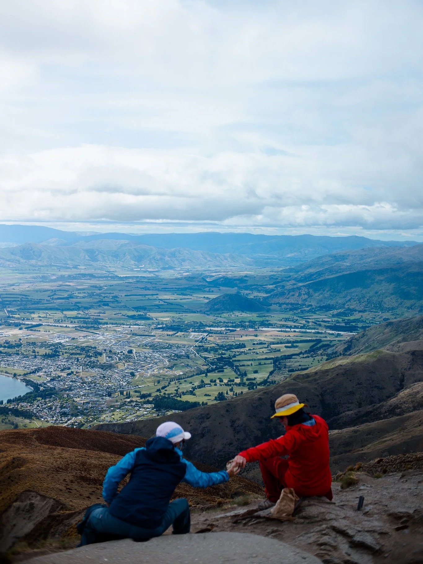 Two children sitting on a mountain trail holding hands with a city and river below and mountains in the distance under cloudy sky.