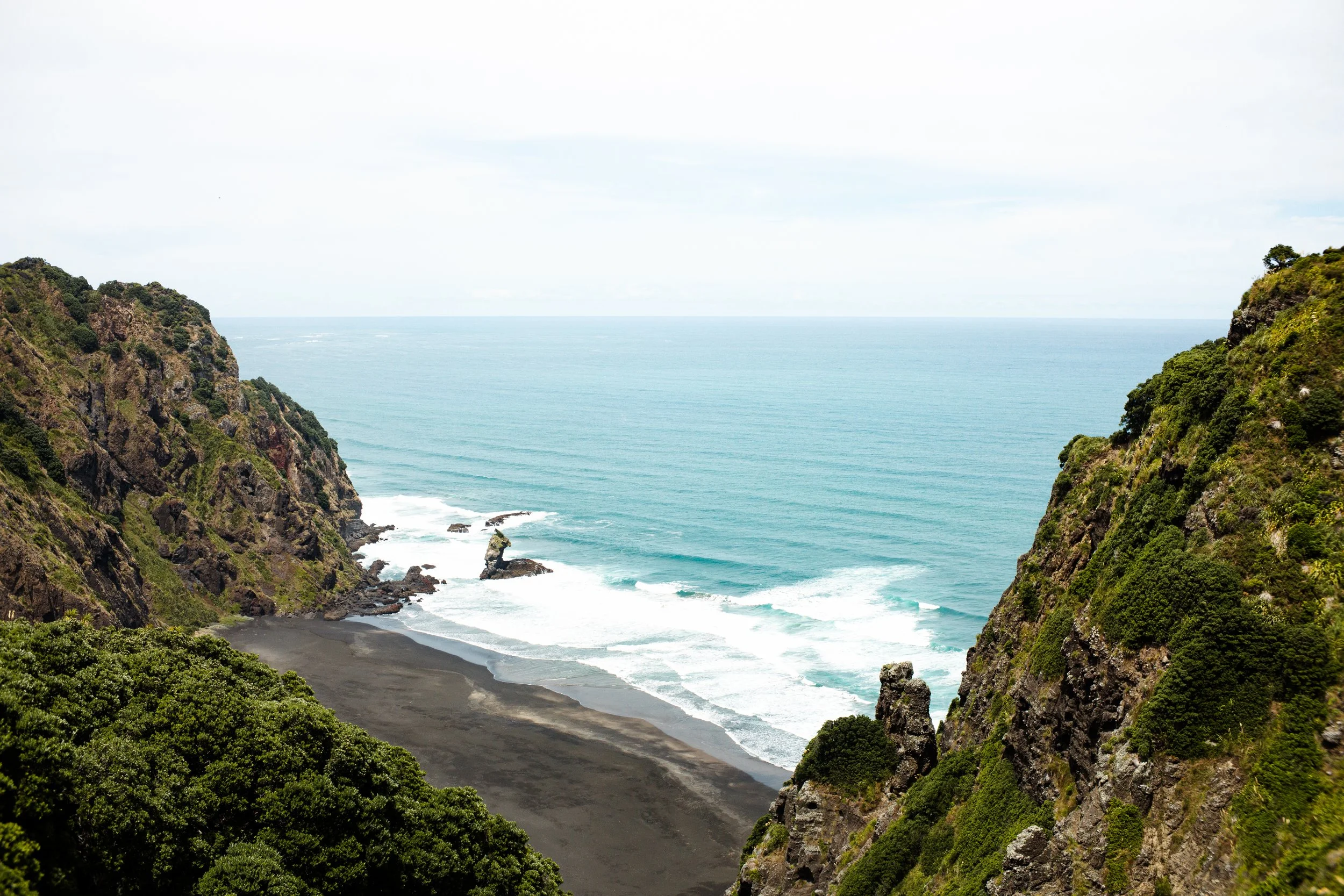 A view of a black sand beach between two green-covered cliffs with the ocean in the background and partly cloudy sky.