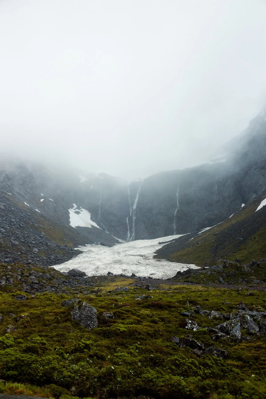 Mountain landscape with patches of snow, waterfalls, and green vegetation in the foreground, shrouded in fog.