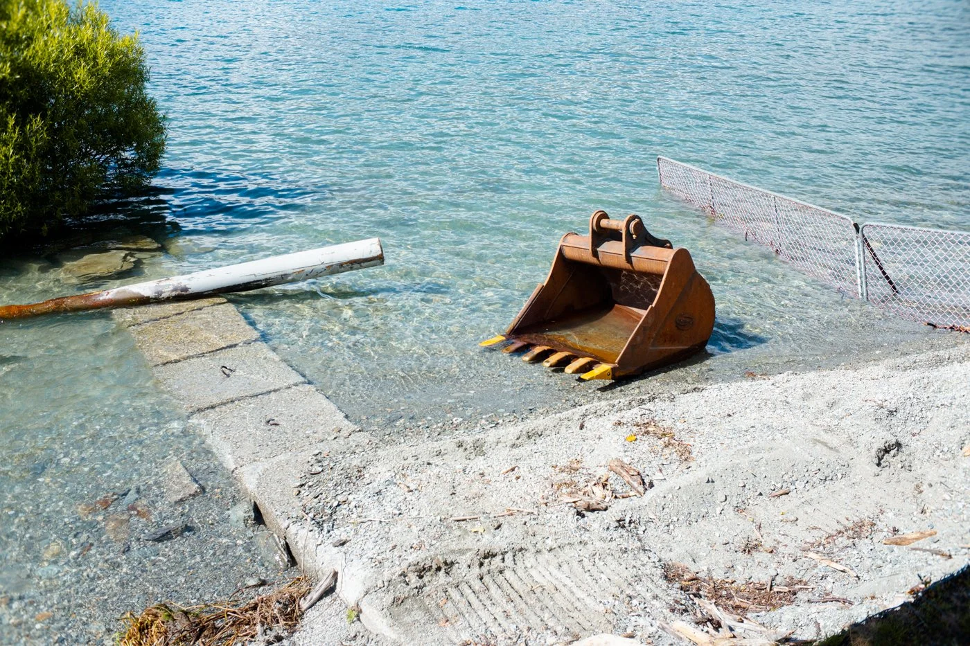 A construction bucket submerged in shallow water near a shoreline, with a white pipeline and orange safety fence partially submerged nearby.