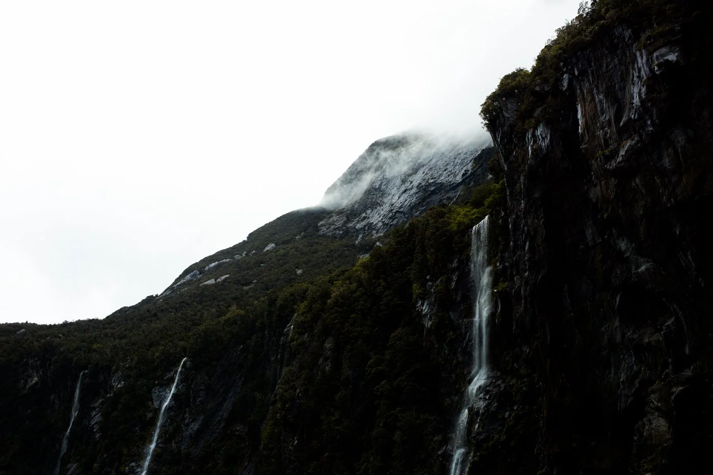 A mountain with a waterfall flowing down its dark, rocky surface, partially covered by fog or low clouds.