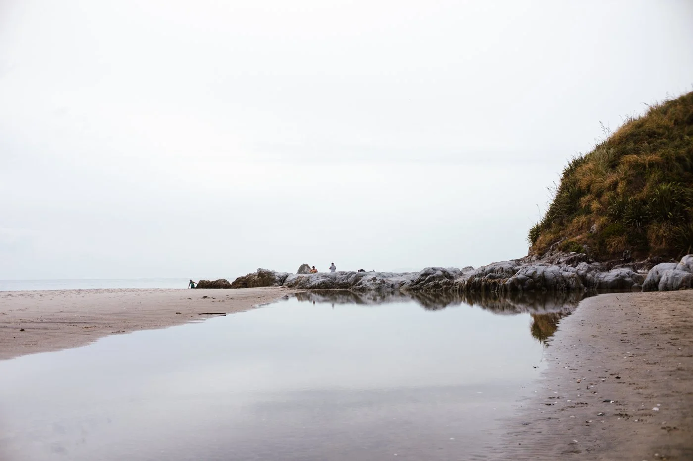 A quiet beach scene with a small creek flowing into the ocean, rocks, and a grassy hill on the right, with a few people visible in the distance.