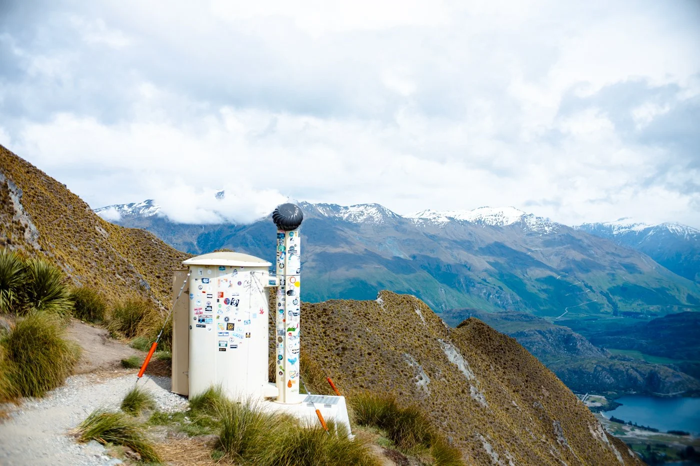 Mountain landscape with a weather station or communication equipment in the foreground, snow-capped mountains in the background, cloudy sky, and a lake in the valley below.