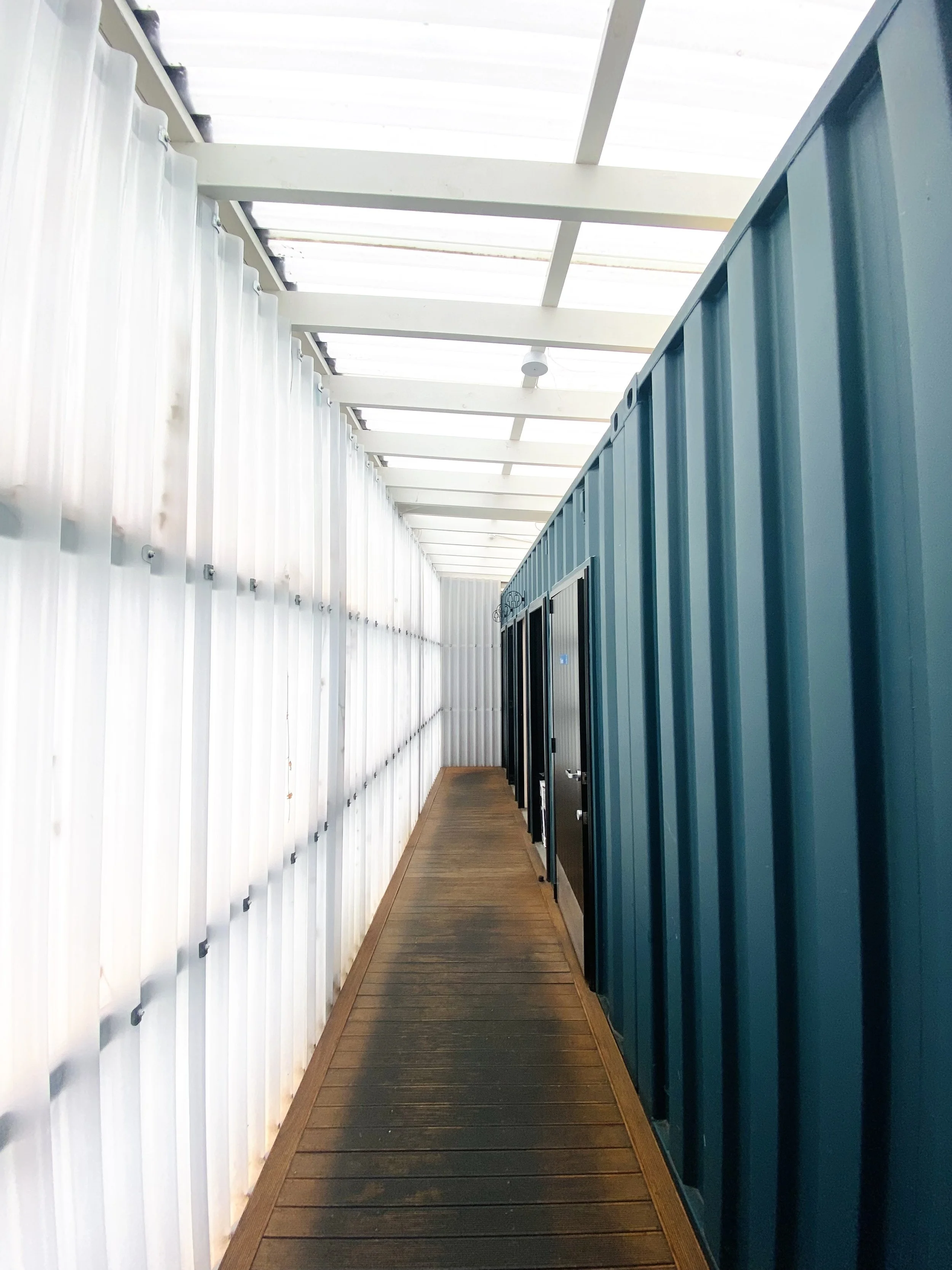 A narrow walkway between white and blue metal storage units with black doors, covered by a white translucent roof.