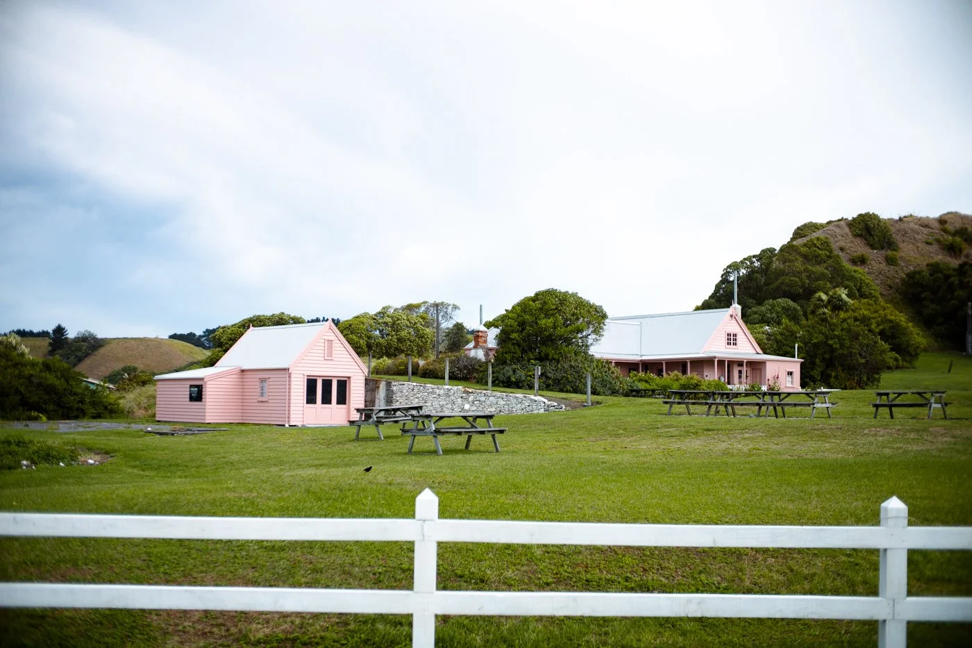 Two pink houses with metal roofs and several picnic tables on a grassy field with a white fence in the foreground, surrounded by green trees and hills under a cloudy sky.