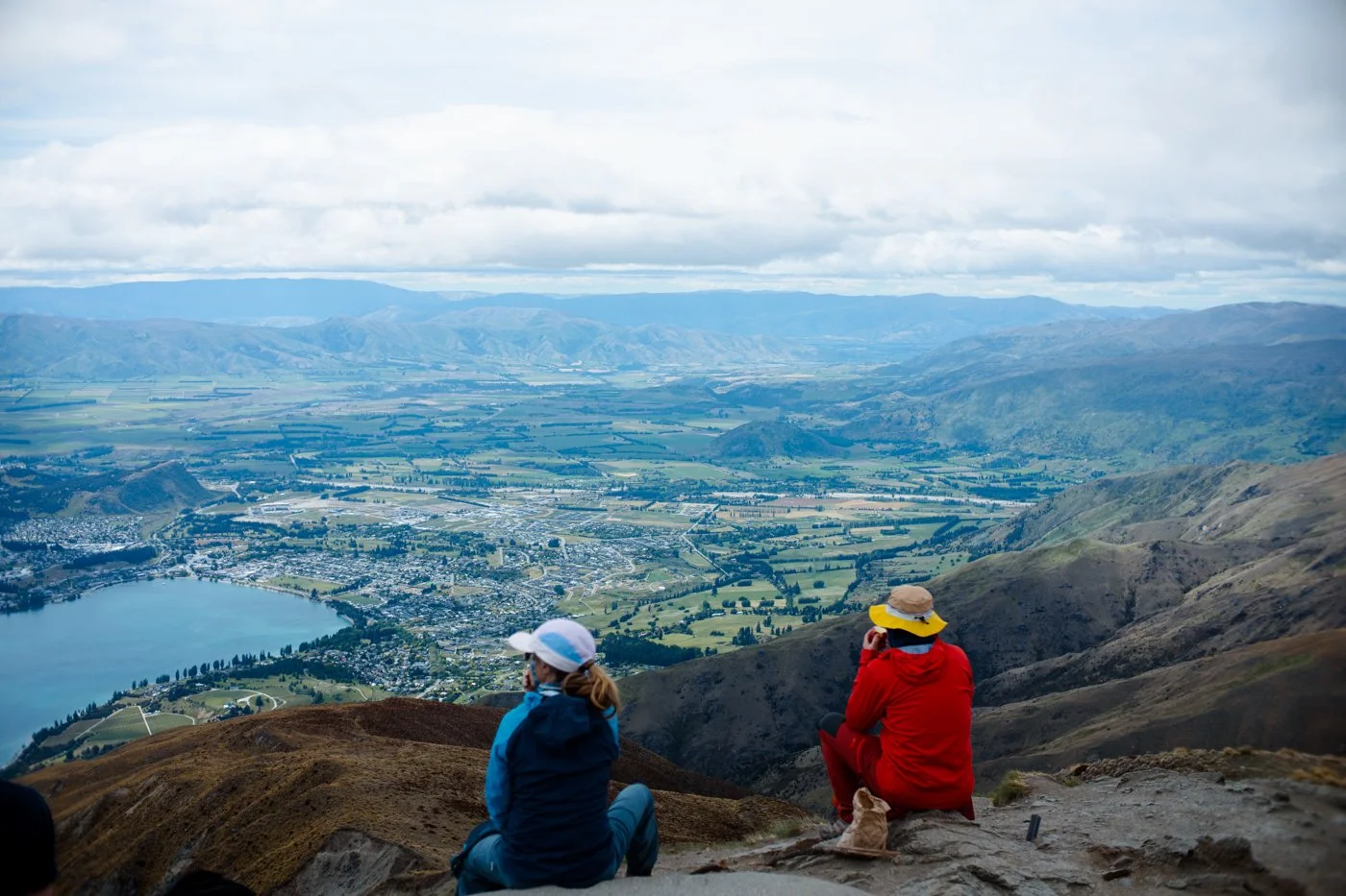 Two hikers sitting on a mountainside overlooking a valley with a lake, town, and surrounding hills in the distance under a cloudy sky.