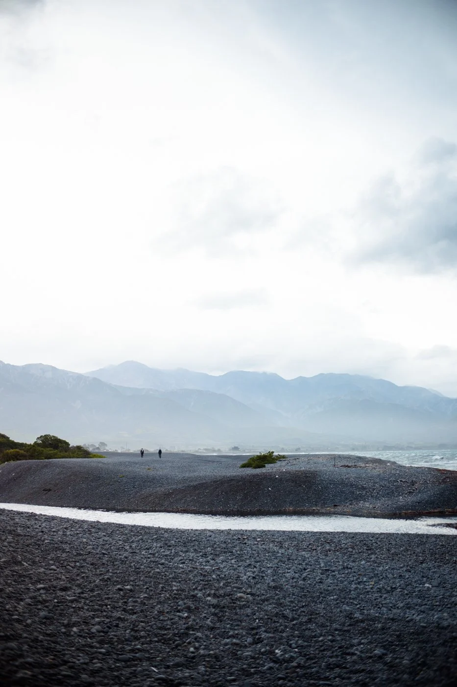 Sea coasts, gravelly shore with a small stream, two distant people, mountain range in the background, cloudy sky.
