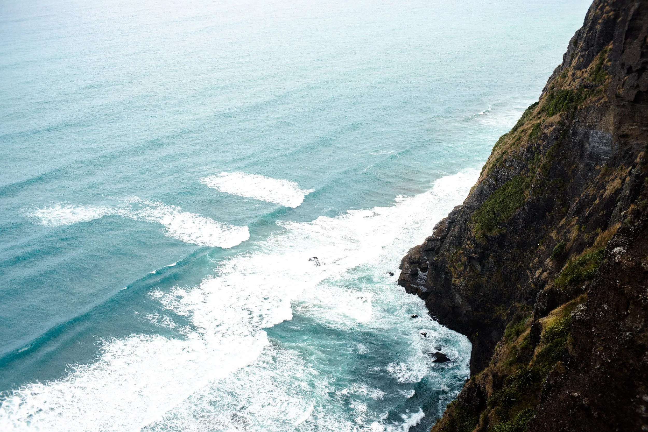 A view of the ocean with waves and a rocky cliffside covered in patches of grass and shrubs.