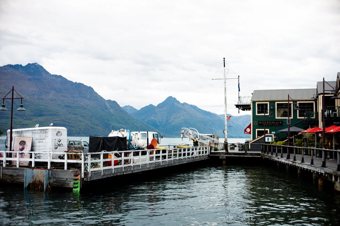 A waterfront pier with buildings, boats, and equipment, set against mountains and cloudy sky in a scenic harbor.