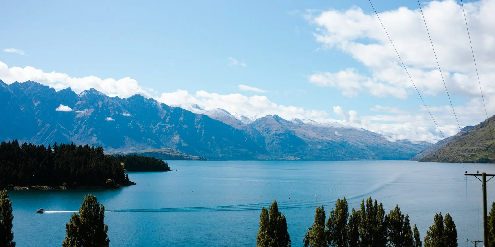 A scenic view of a large lake surrounded by mountains with snow-capped peaks, green forested areas, and a boat creating a wake in the water.