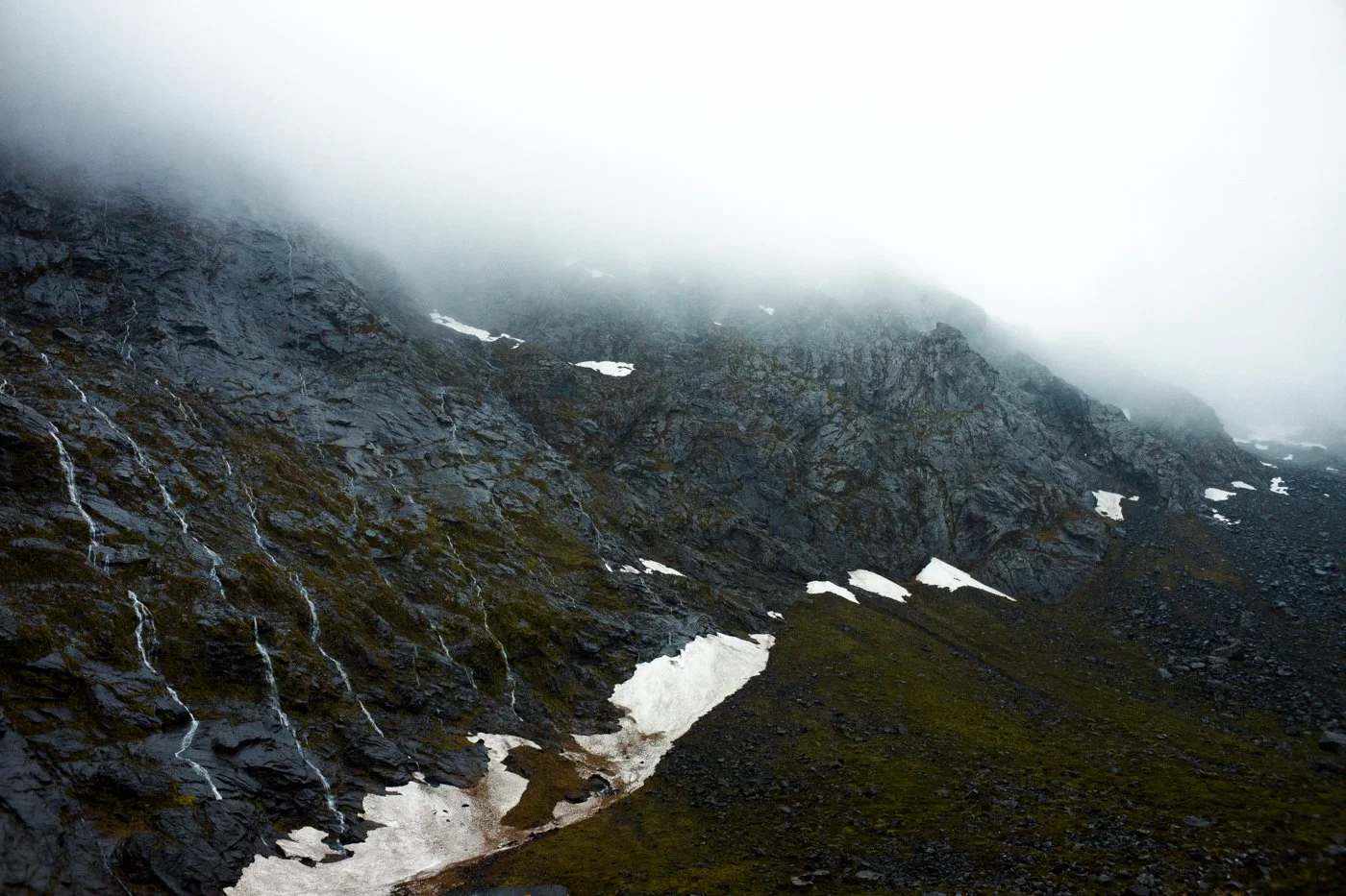 A mountain landscape with dark rock formations, patches of snow, and a misty sky overhead.