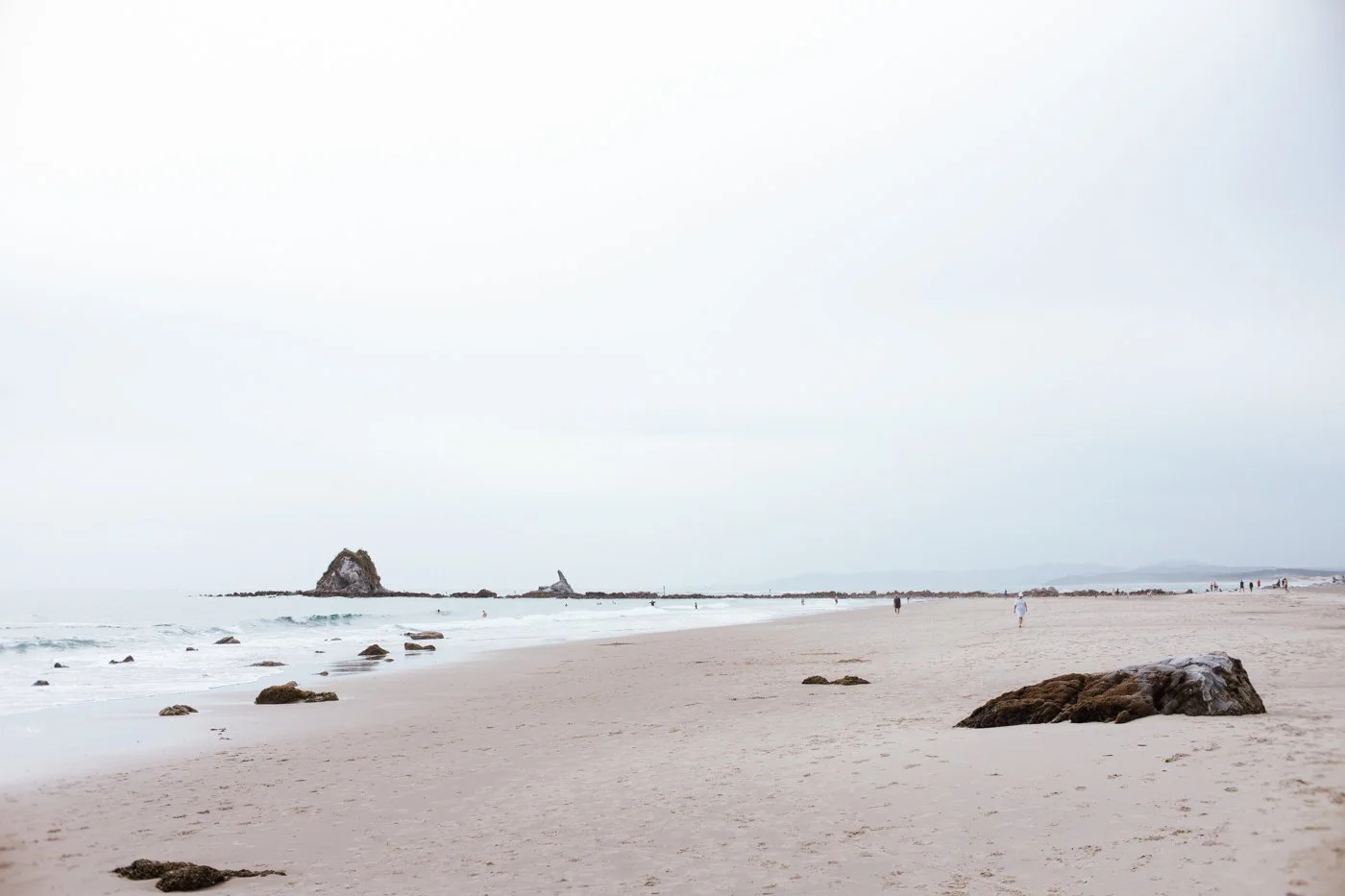 A sandy beach with rocks scattered along the shore, a few people walking, and rock formations in the distance under an overcast sky.