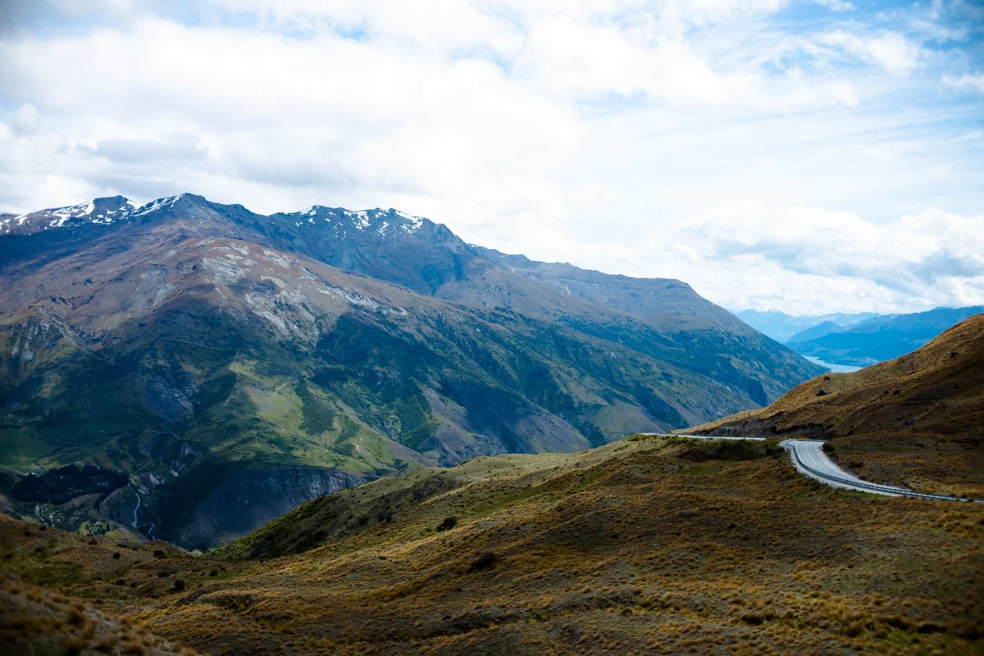 Scenic mountain landscape with winding road, green and brown hills, and snow-capped peaks in the distance under a cloudy sky.