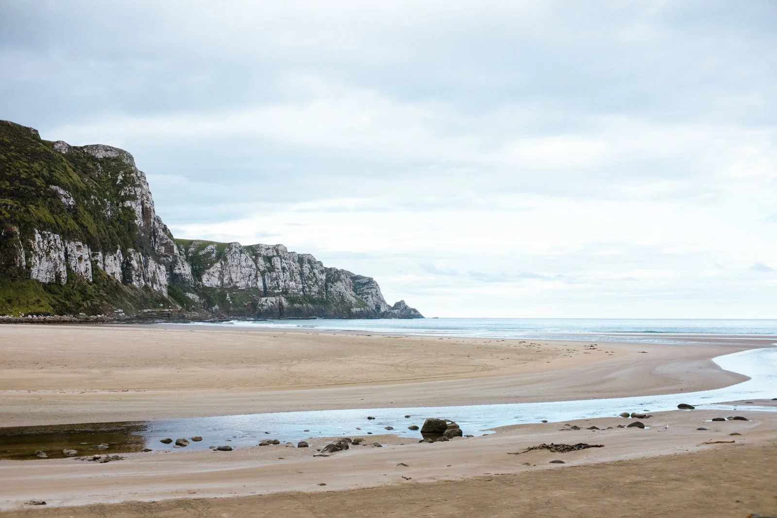 A serene beach with large cliffs on the left, sandy shores, and a gentle stream flowing across the sand under a cloudy sky.