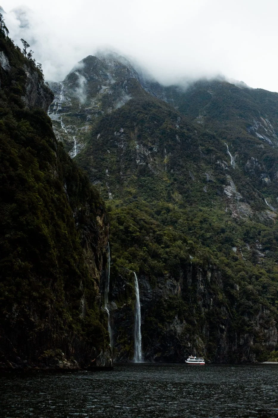 A mountainous landscape with clouds and waterfalls falling down the cliffs into a body of water, with a boat near the base of the waterfalls.