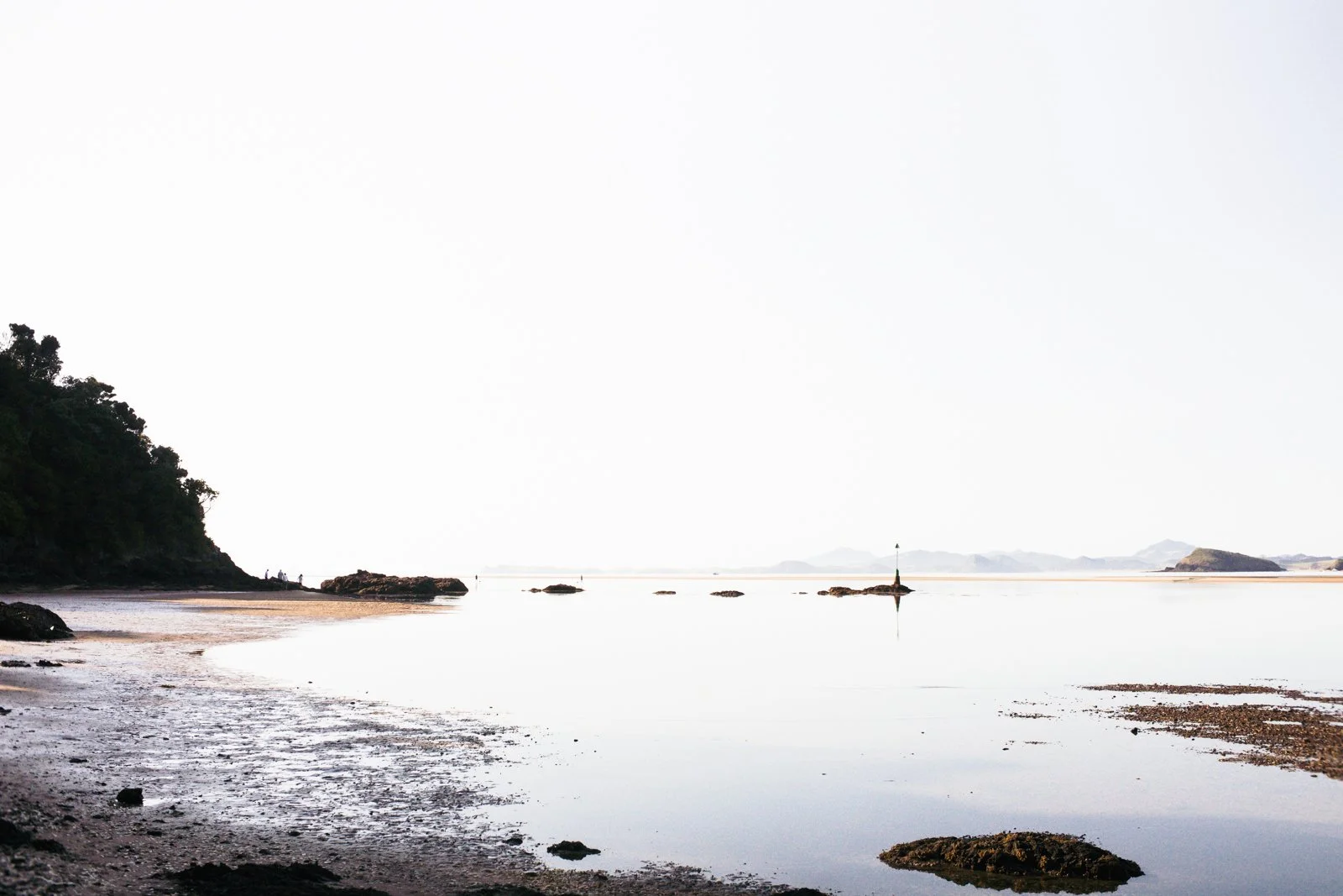 Calm coastal scene with a sandy shore, rocks, a small island or landmass in the background, and a navigation marker on a rock outcrop, under a bright, clear sky.