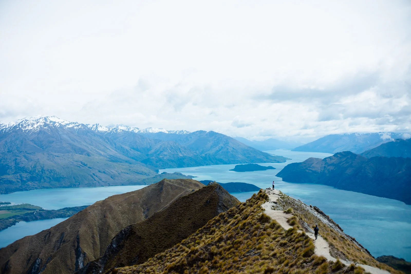 A hiker standing on a mountain ridge overlooking a lake with mountain ranges in the background.