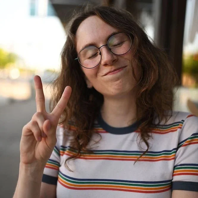 A young woman with curly brown hair wearing glasses and a striped shirt, smiling with her eyes closed and making a peace sign with her right hand.