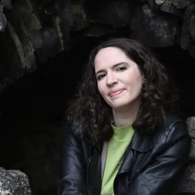 A young woman with curly dark hair and fair skin, wearing a black leather jacket and a lime green shirt, is sitting outdoors near rocks and a cave or tunnel opening.