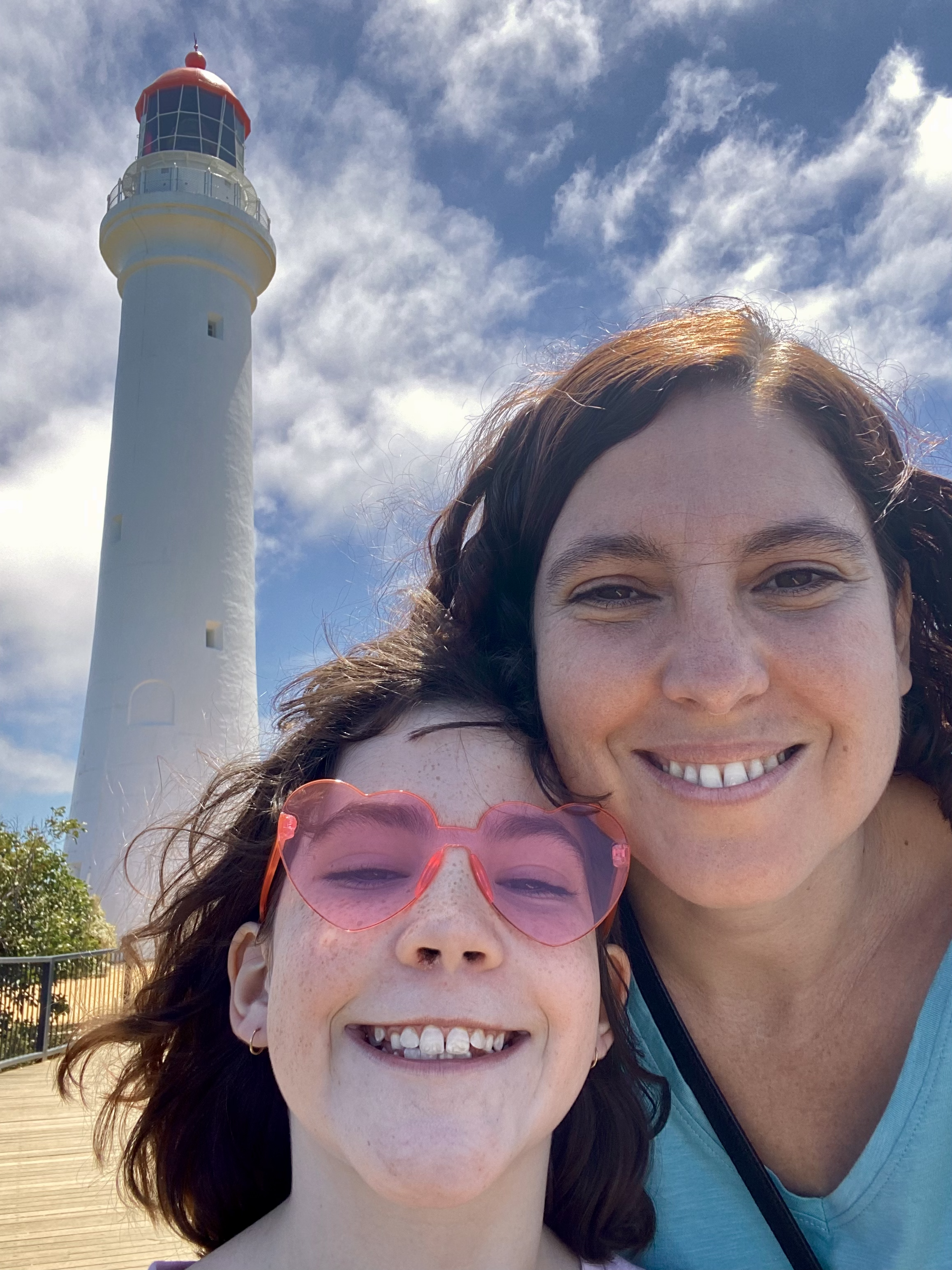 A woman and a girl smiling for a selfie near a lighthouse on a sunny day with a partly cloudy sky.