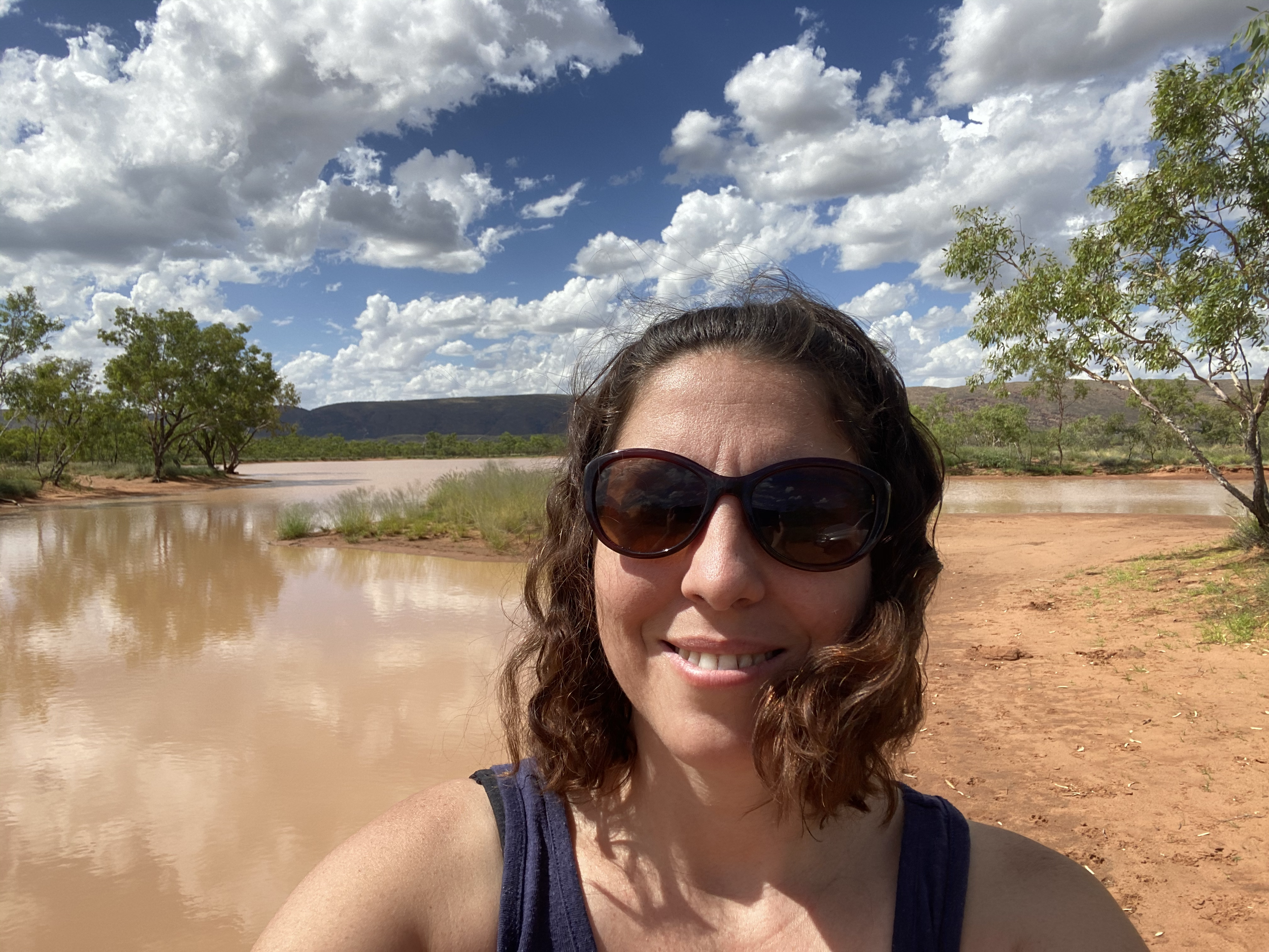 Woman with curly hair wearing sunglasses and a navy tank top taking a selfie near a river or lake with a muddy bank, green trees, mountains in the distance, and a partly cloudy sky.