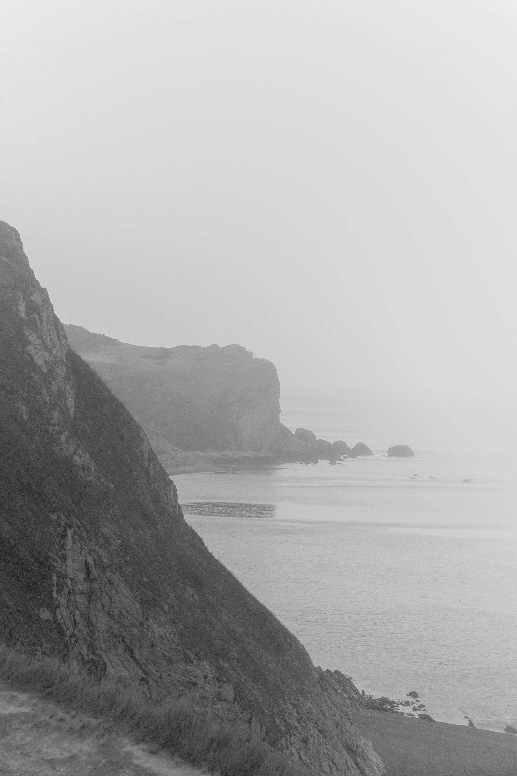 Black and white photo of rugged coastal cliffs with ocean waves and rocks in the distance under a foggy sky.