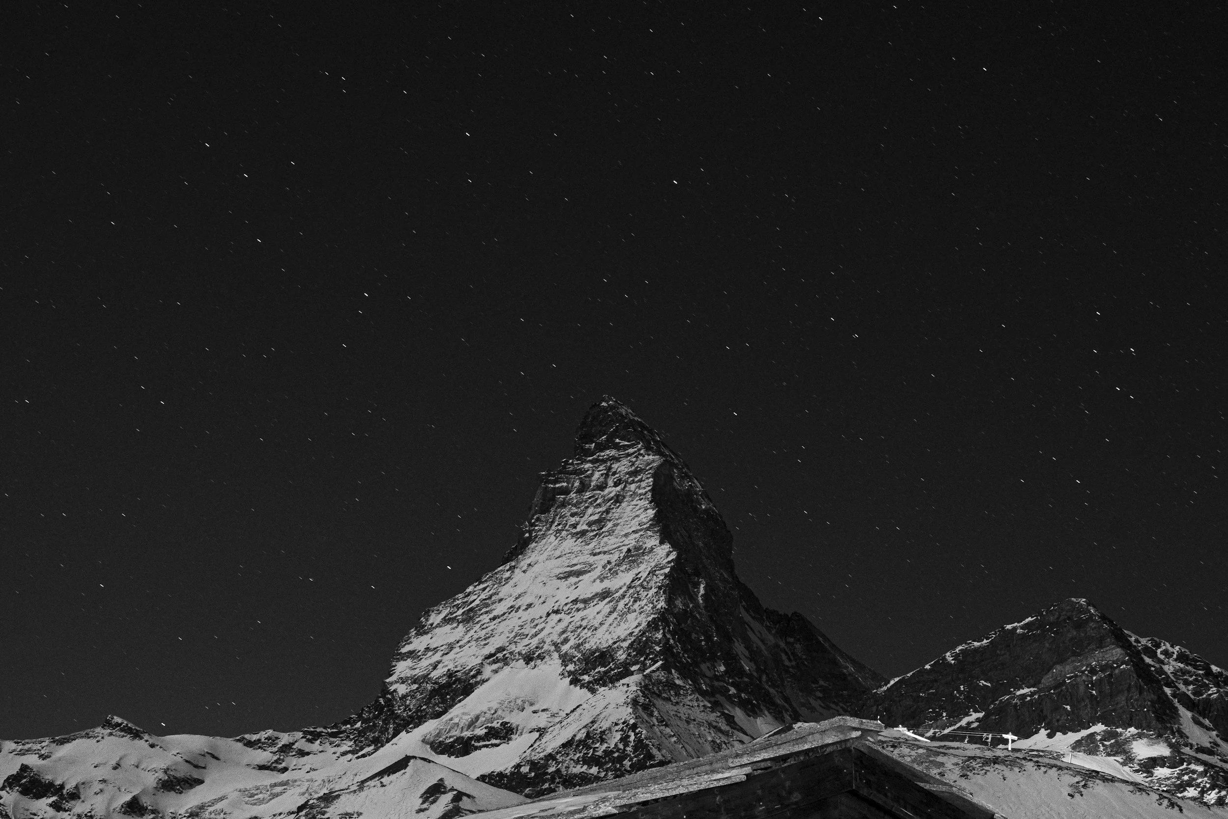 Nighttime view of snowy mountain peak, possibly the Matterhorn, under a starry sky.