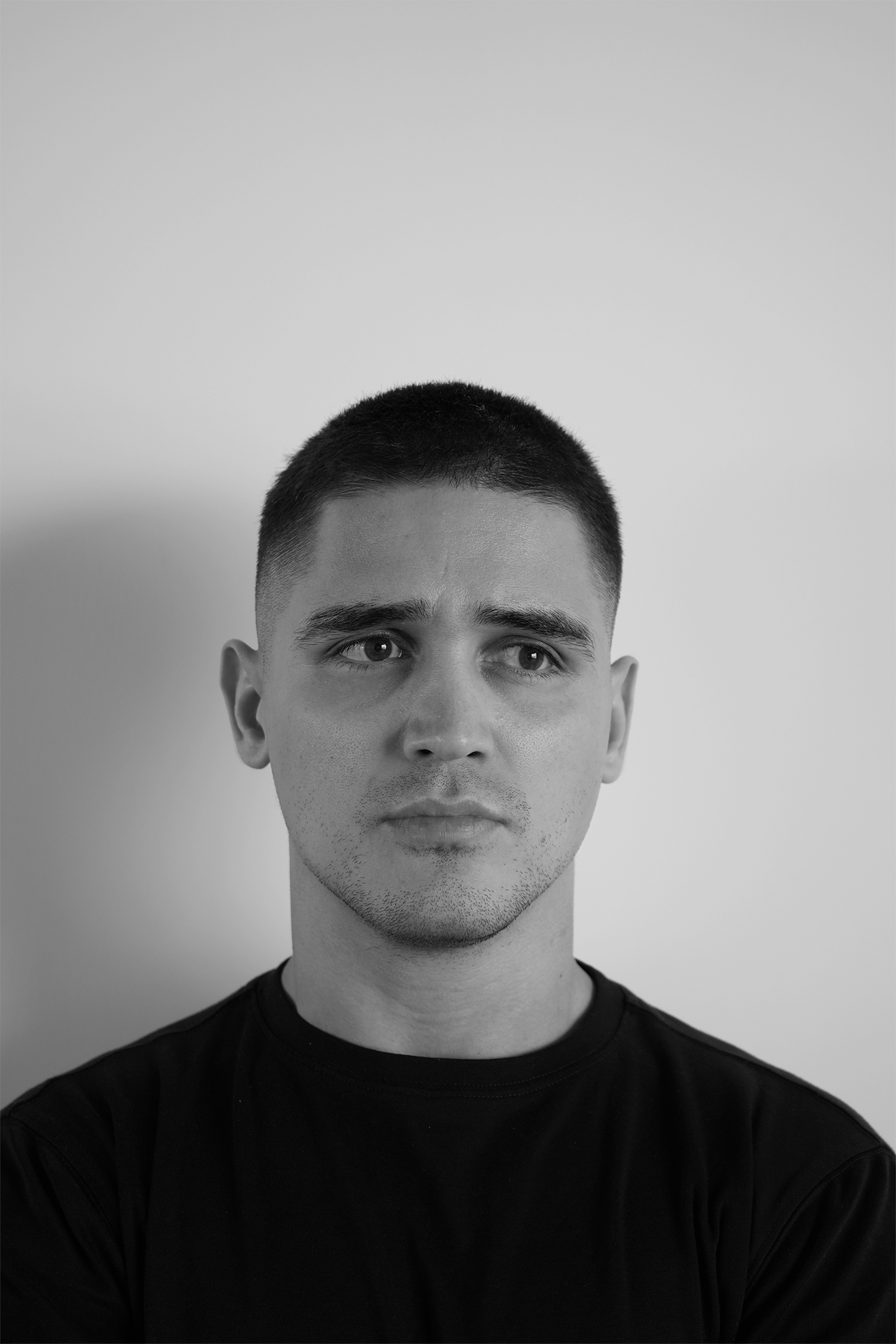 Black and white portrait of a young man with short hair, wearing a black shirt, looking directly at the camera with a neutral expression.