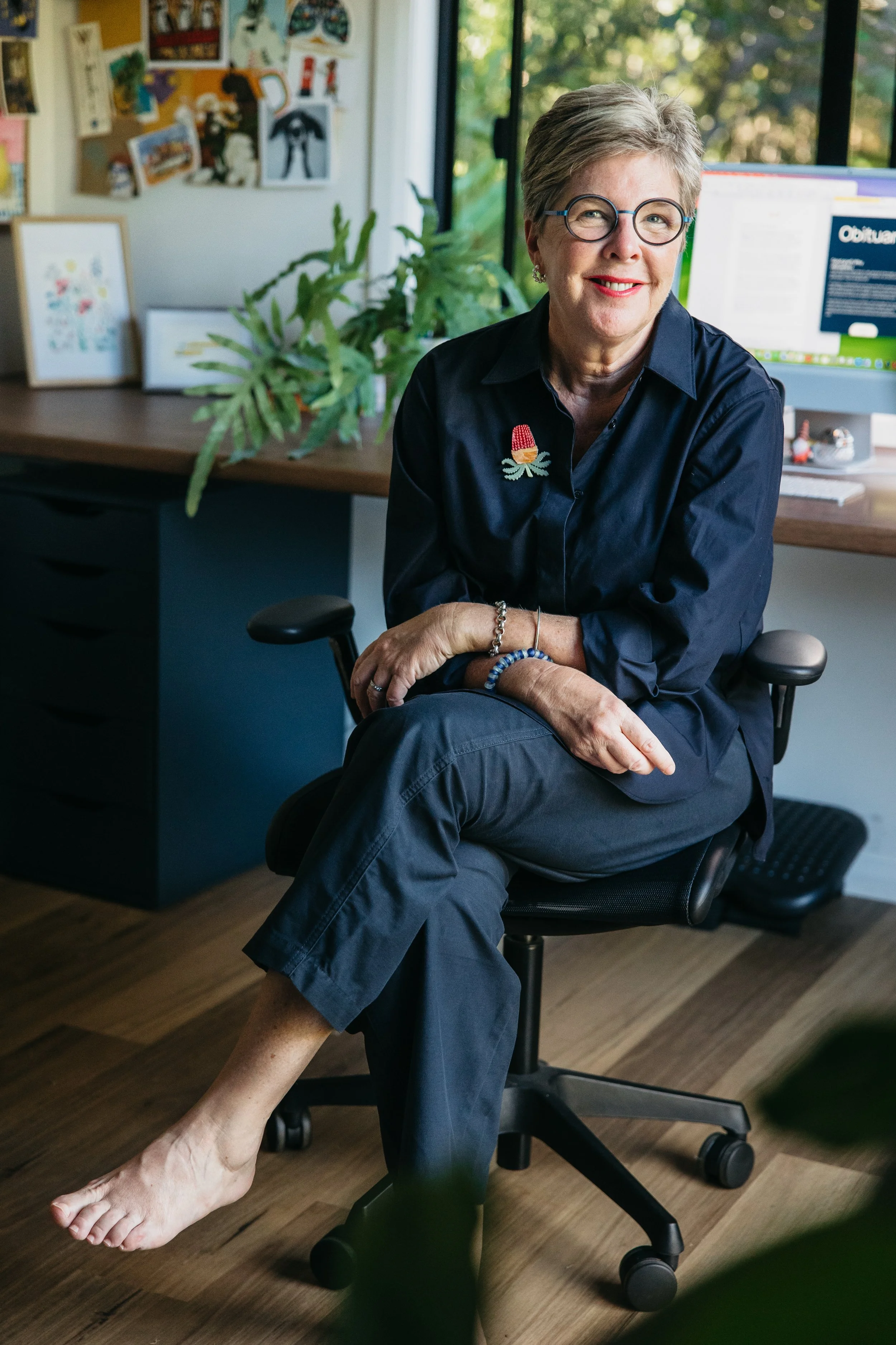 Woman in dark shirt, wearing glasses, sits at desk with plant and computer