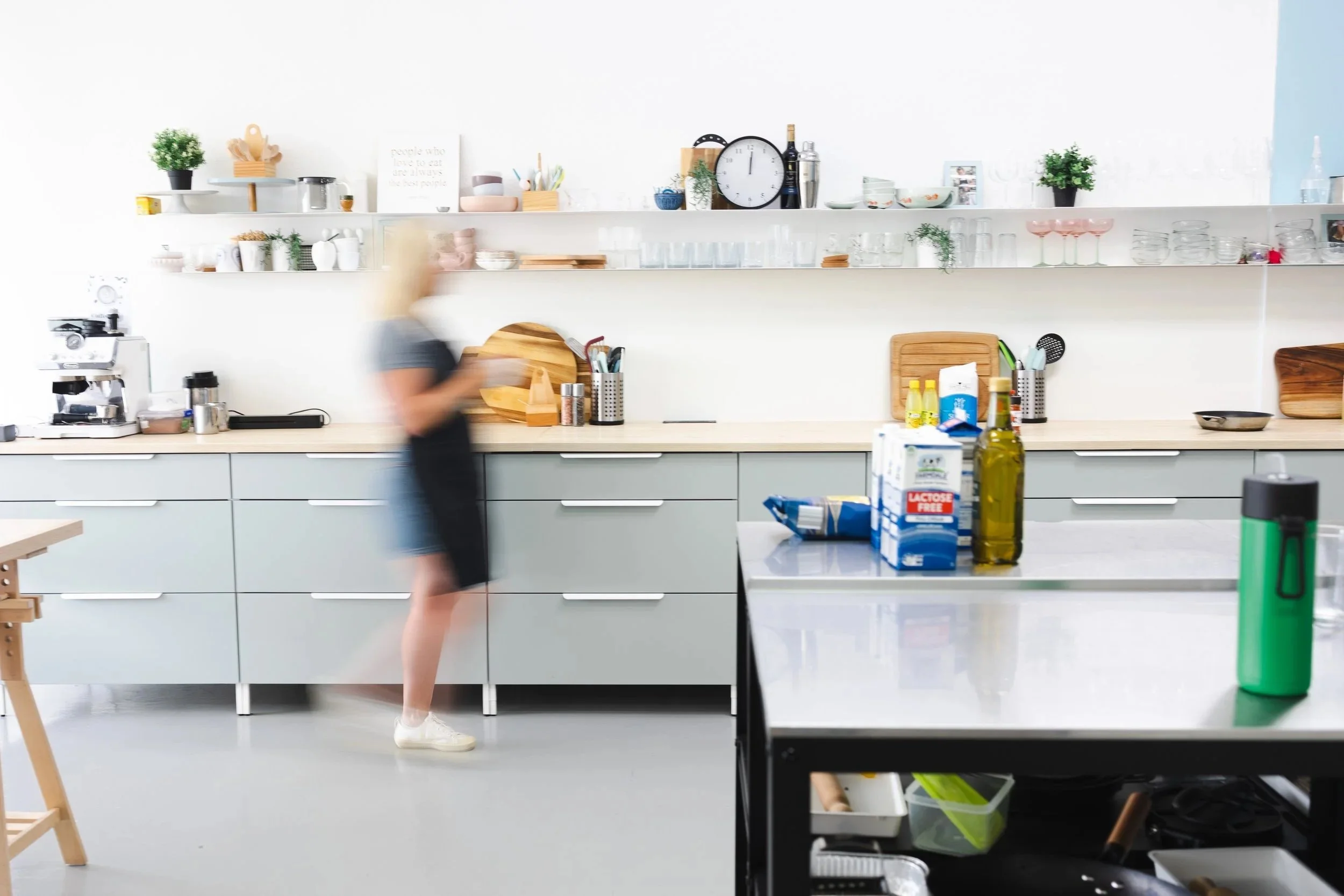 Blurred person moving in a modern kitchen with grey cabinets, open shelves, and various kitchen items on the countertops and shelves.