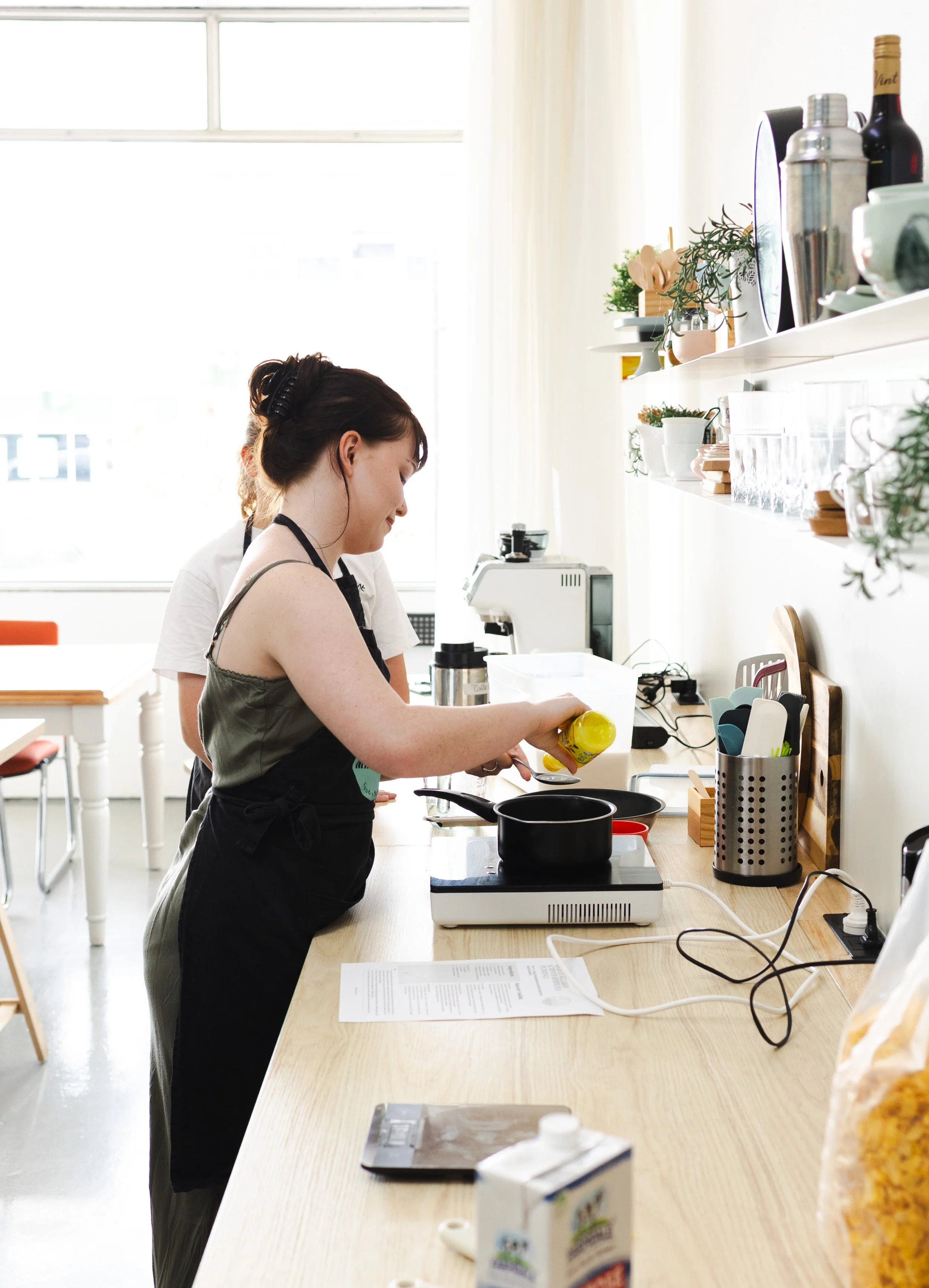 A woman in a black apron cooking on a stove in a bright kitchen, with another person partially visible behind her, on a kitchen counter with utensils, plants, and bottles, near a window with white curtains.