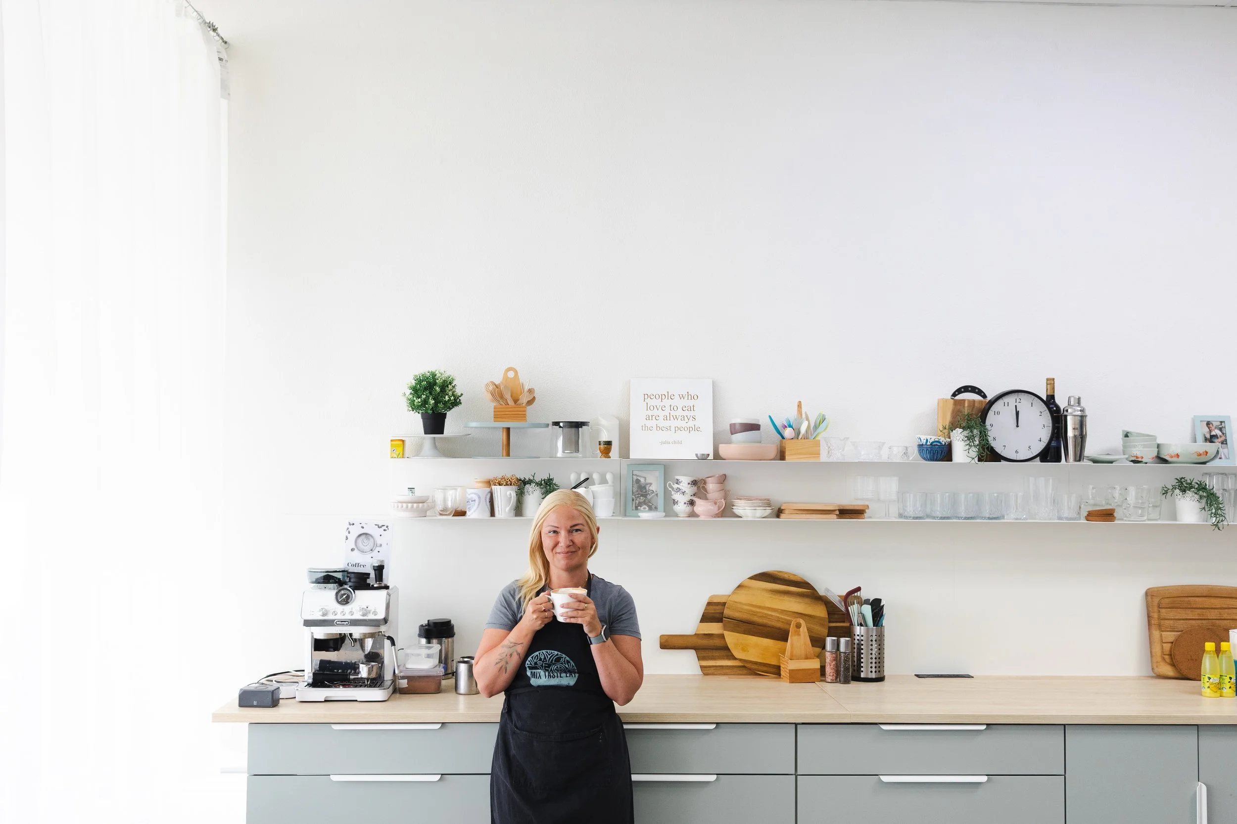 A woman with blonde hair smiling and holding a mug in a modern kitchen with white walls, wooden countertops, and open shelves with dishes, plants, and kitchen utensils.