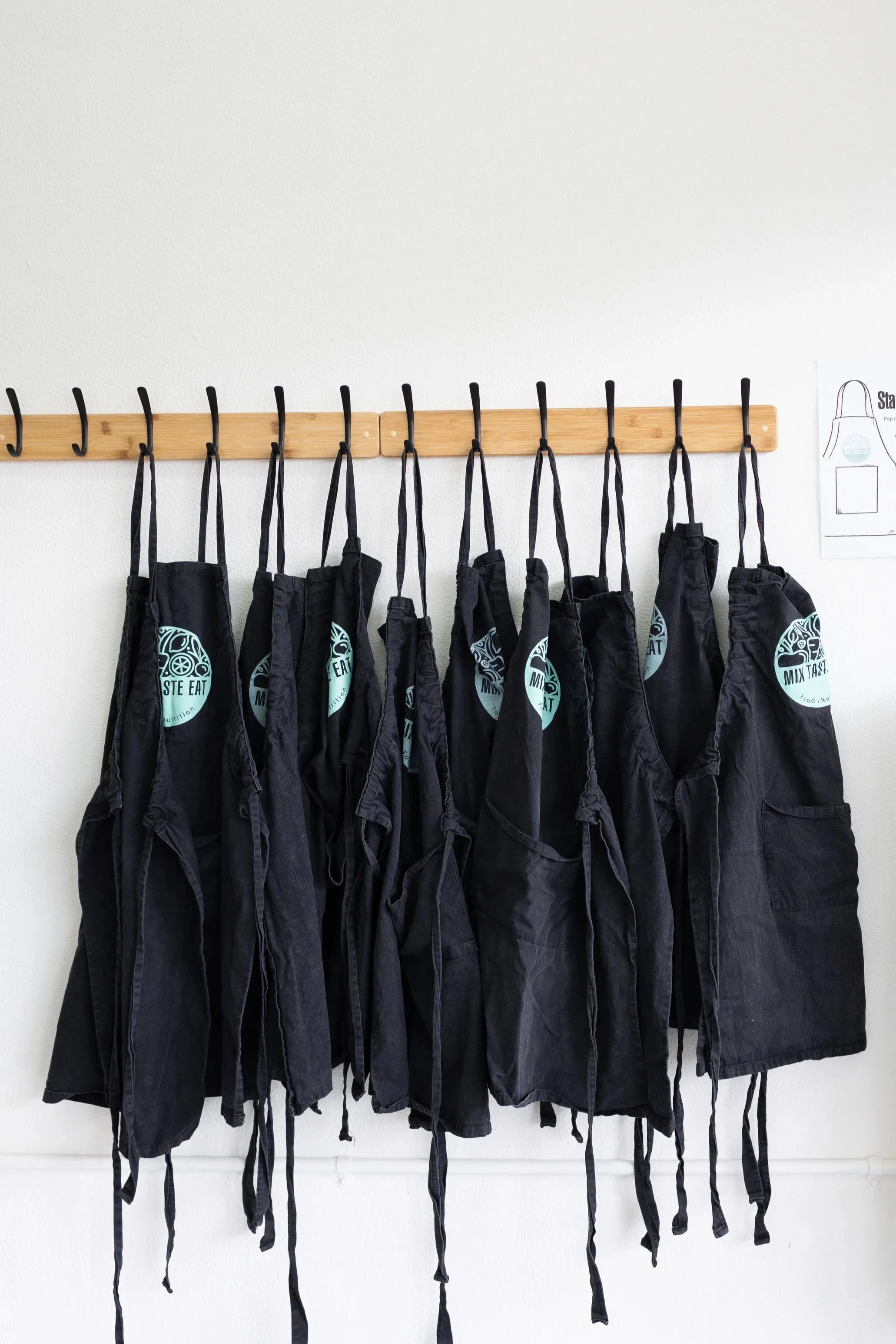 Nine black aprons hanging on a wooden rack against a white wall.