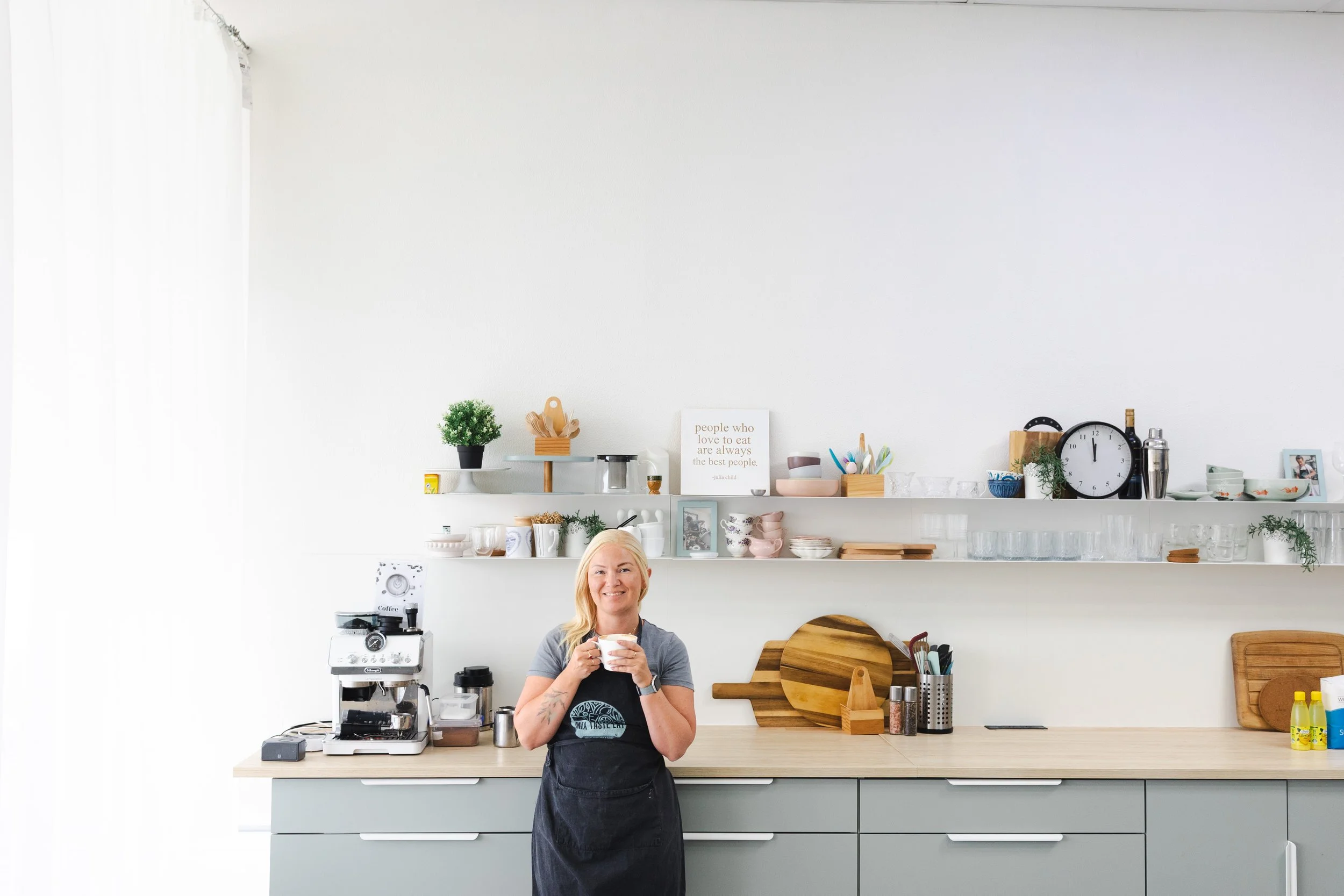 A woman with blonde hair smiling and holding a mug stands in a modern, minimalist kitchen. The kitchen features a light wooden countertop, gray cabinets, and a white wall with two white shelves holding kitchenware, a clock, plants, and decorative items.