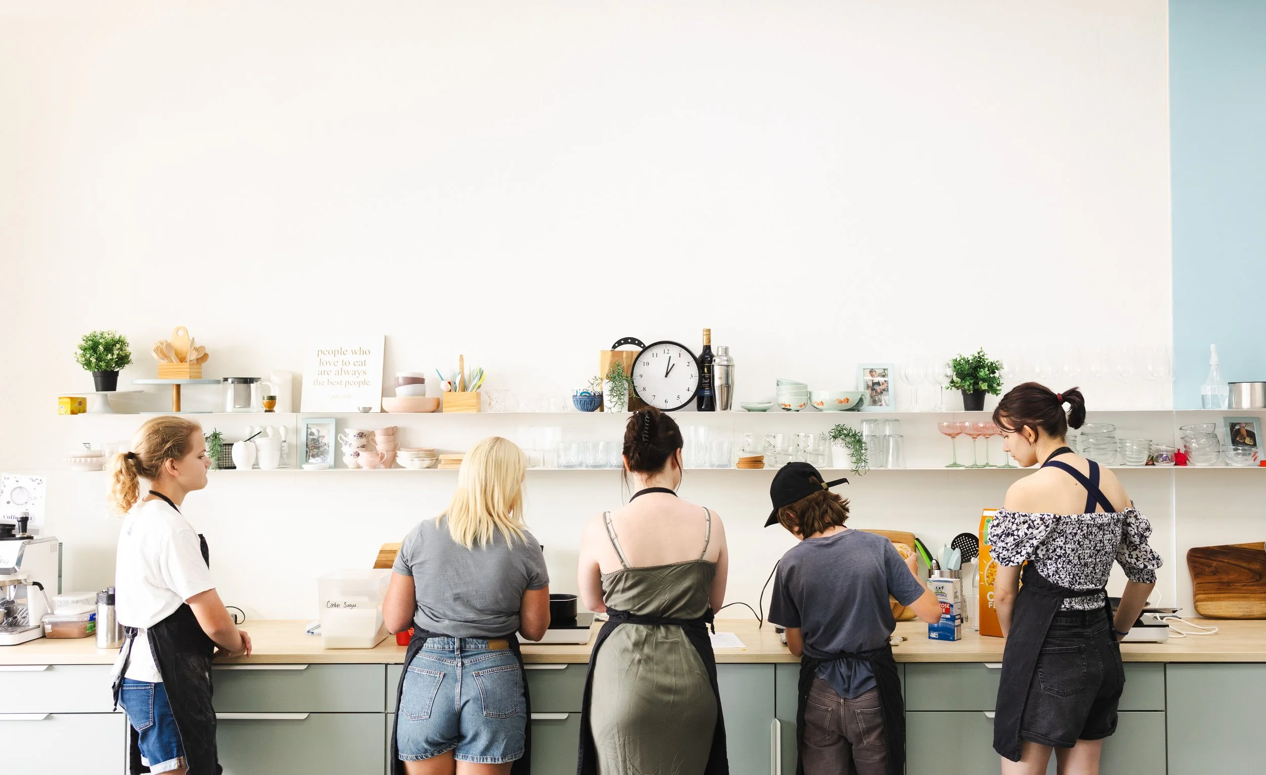 Five young women and girls preparing food in a modern kitchen with white and green cabinets, open shelves with dishes, glasses, and decorative items, and a large wall clock.