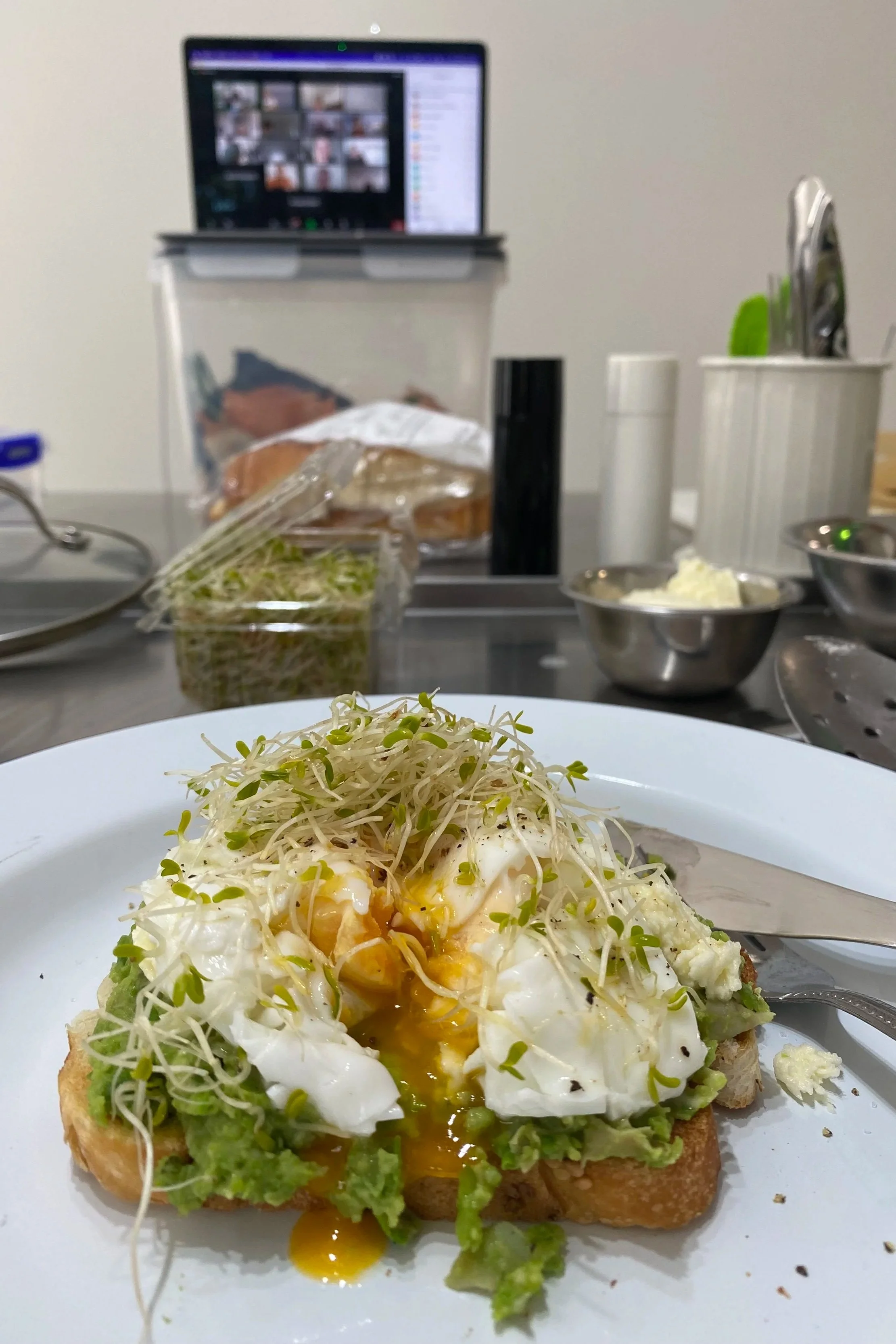 A plate with avocado toast topped with a soft boiled egg and alfalfa sprouts, on a kitchen counter with various bowls, utensils, and a laptop in the background.