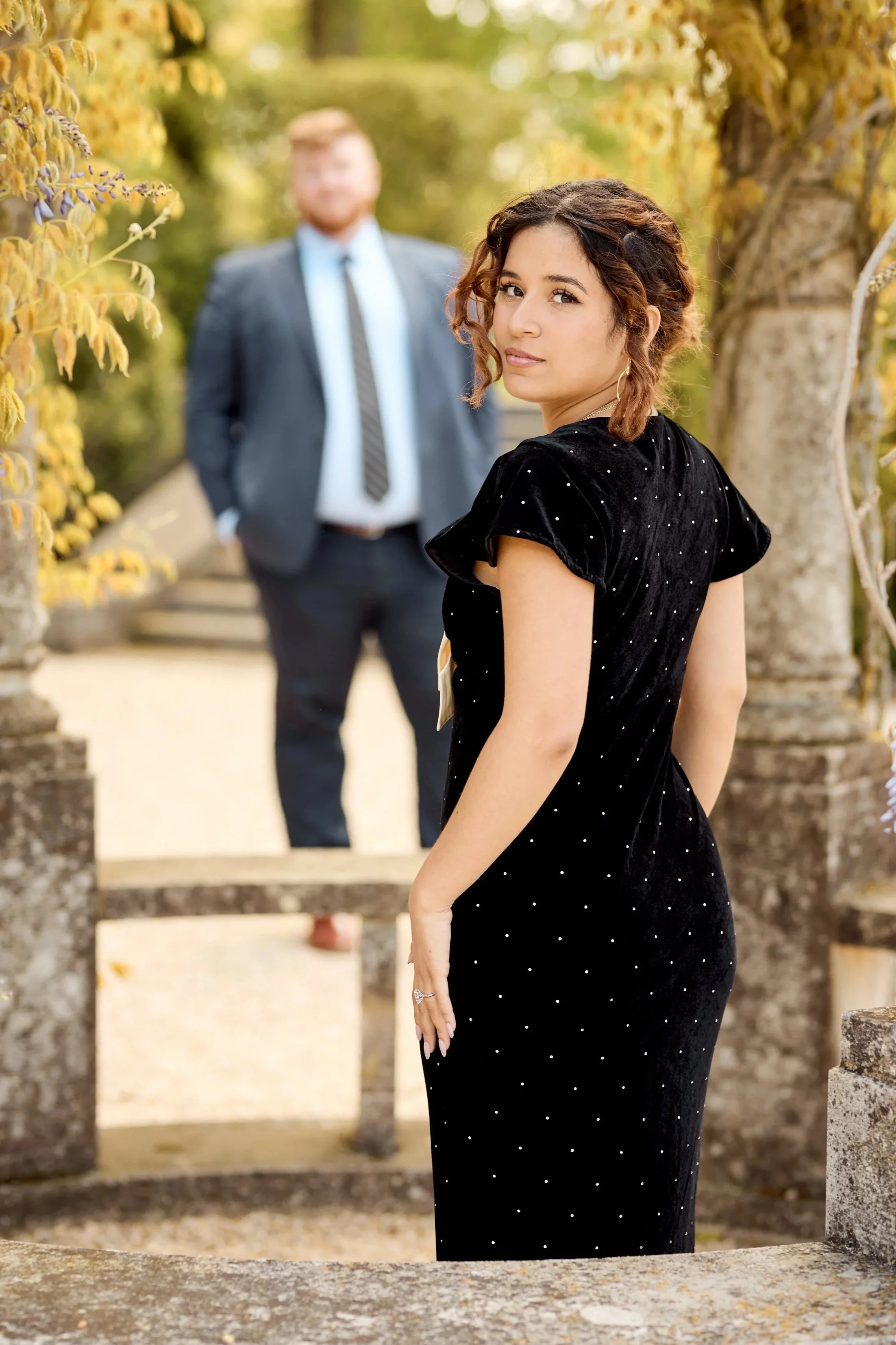 A woman with curly brown hair and a black dress with small white dots is standing outdoors surrounded by yellow autumn leaves. Behind her, a man in a suit and tie is slightly out of focus.